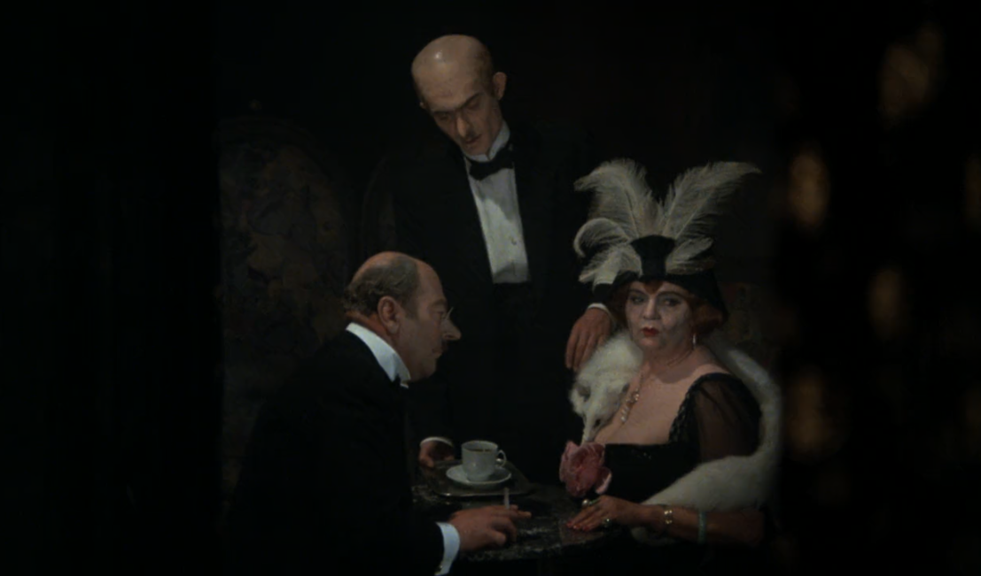 Zdjęcie przedstawia kobietę w czarnej sukni otuloną białym futrem. Ma na głowie kapelusz z białymi piórami. Siedzi przy stoliku z mężczyzną w garniturze. Mężczyzna pali papierosa. Na stoliku stoi filiżanka. Za stolikiem stoi łysy mężczyzna w garniturze.