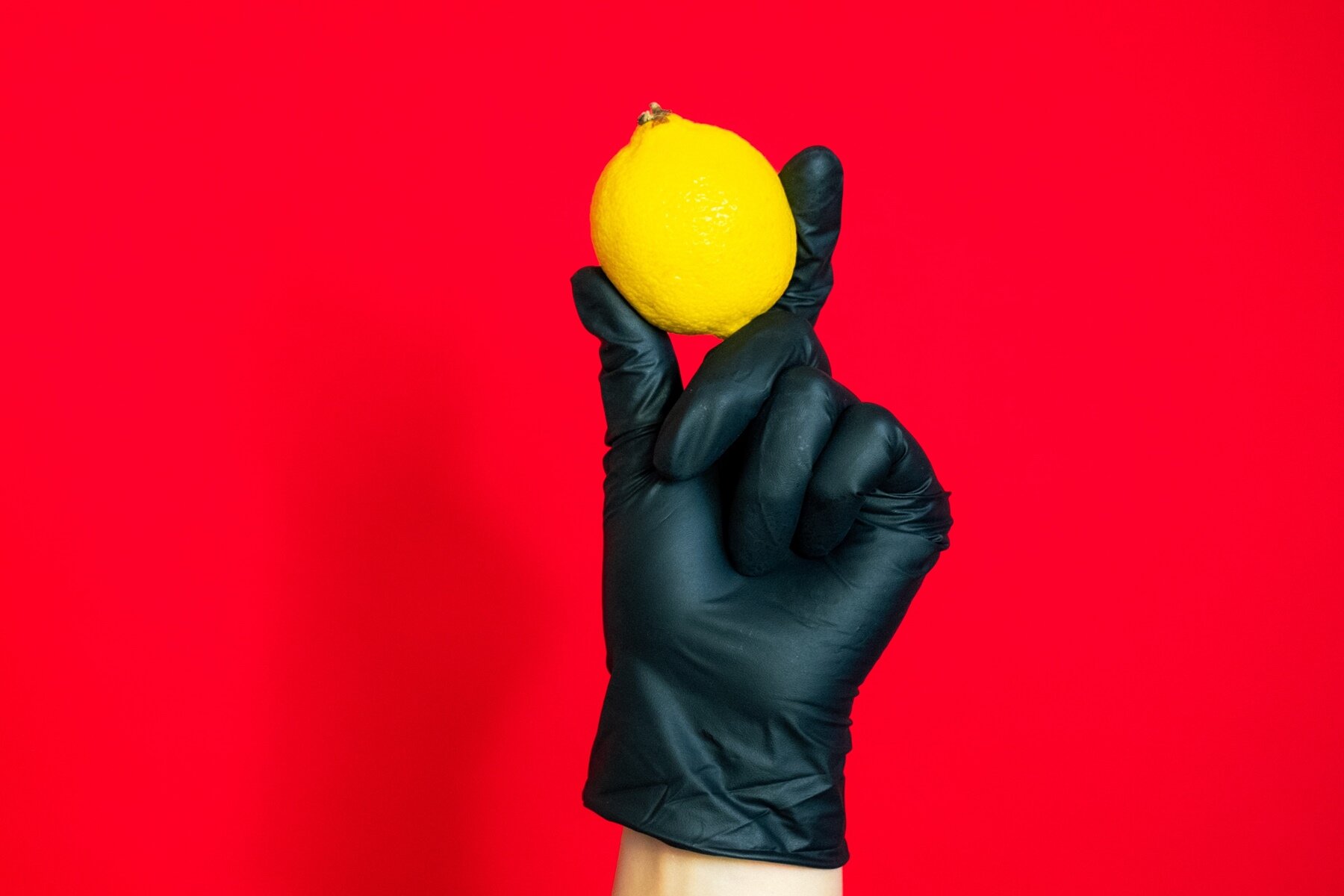 Zdjęcie przedstawia lewą dłoń w jednorazowej, czarnej lateksowej rękawiczce. Kciuk i palec wskazującym dłoni przytrzymują żółtą cytrynę. Pozostałe palce są zgięte. Tło zdjęcia jest czerwone. 