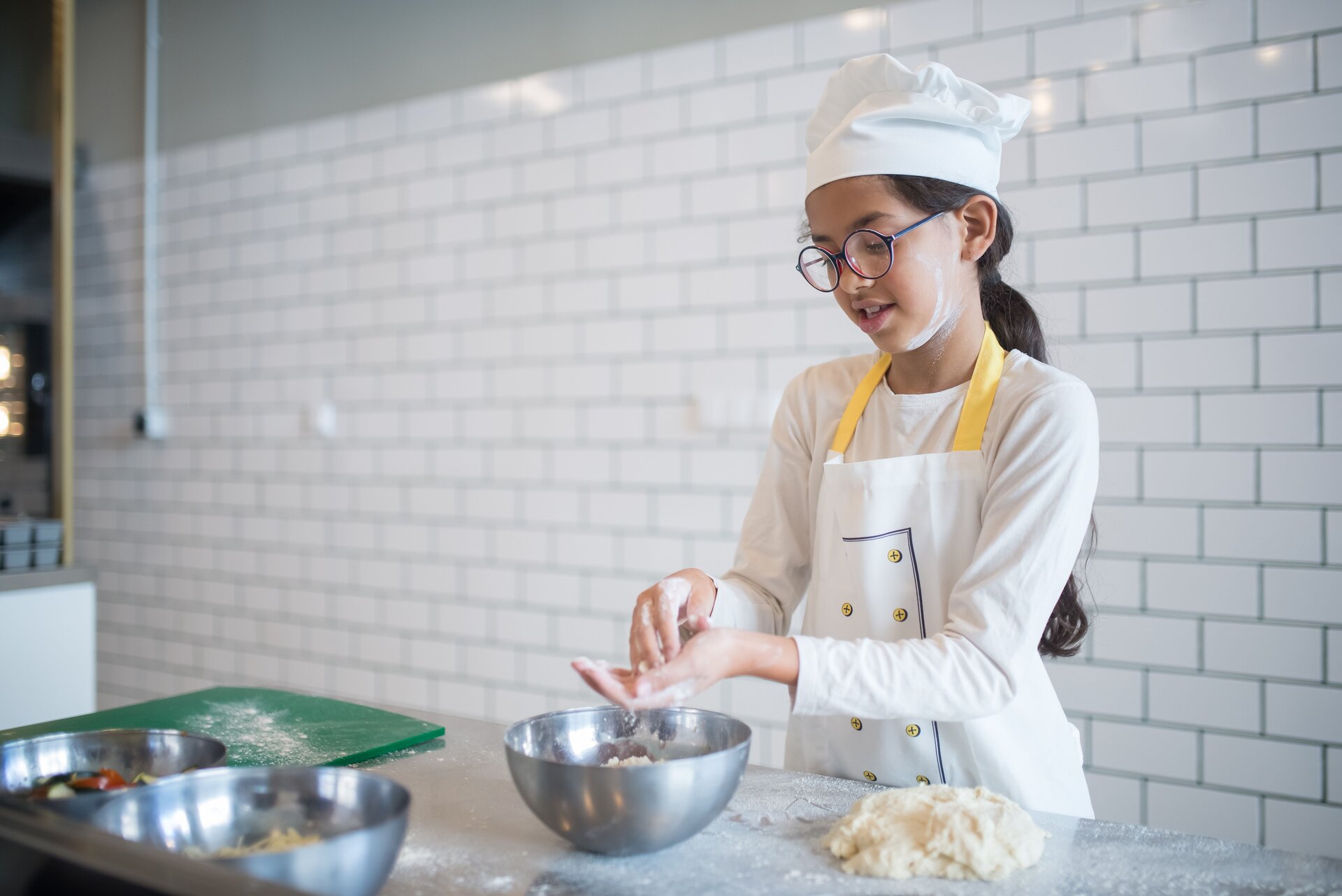 Zdjęcie przedstawia dziewczynkę stojącą przy blacie kuchennym. Dziewczynka ma na sobie fartuszek oraz nakrycie głowy kucharza. Długie ciemne włosy ma upięte z tyłu. Jej buzia jest ubrudzona mąką. Dziewczynka ma lekko ugięte ręce, które trzyma nad metalową miską stojącą na blacie. W dłoniach przesypuje mąkę. Na prawo na posypanym mąką blacie widoczne jest przygotowane, zagniecione ciasto. Z przodu widoczne są metalowe miski oraz zielona deska do krojenia.