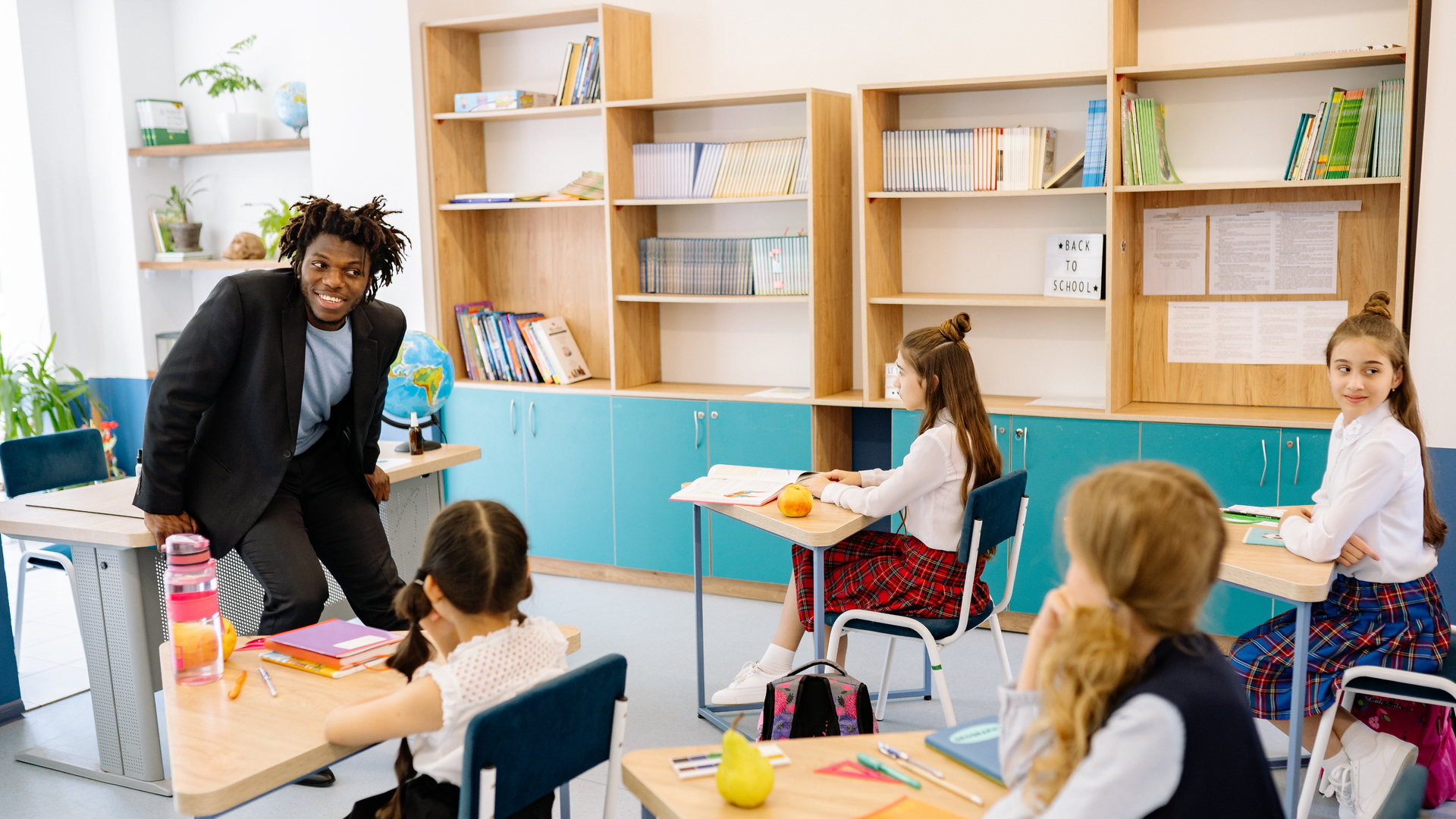 Zdjęcie przedstawia salę lekcyjną. W ławkach siedzą uczniowie. Na zdjęciu widoczne są cztery dziewczynki. Każda z nich siedzi na krześle w swojej ławce. Przed nimi, oparty o biurko stoi uśmiechnięty nauczyciel z krótkimi dredami na głowie. Za biurkiem stoi krzesło. Na drugim planie przy ścianie widoczne są regały na książki oraz półki. Na półkach i na parapecie znajdują się rośliny w doniczkach.