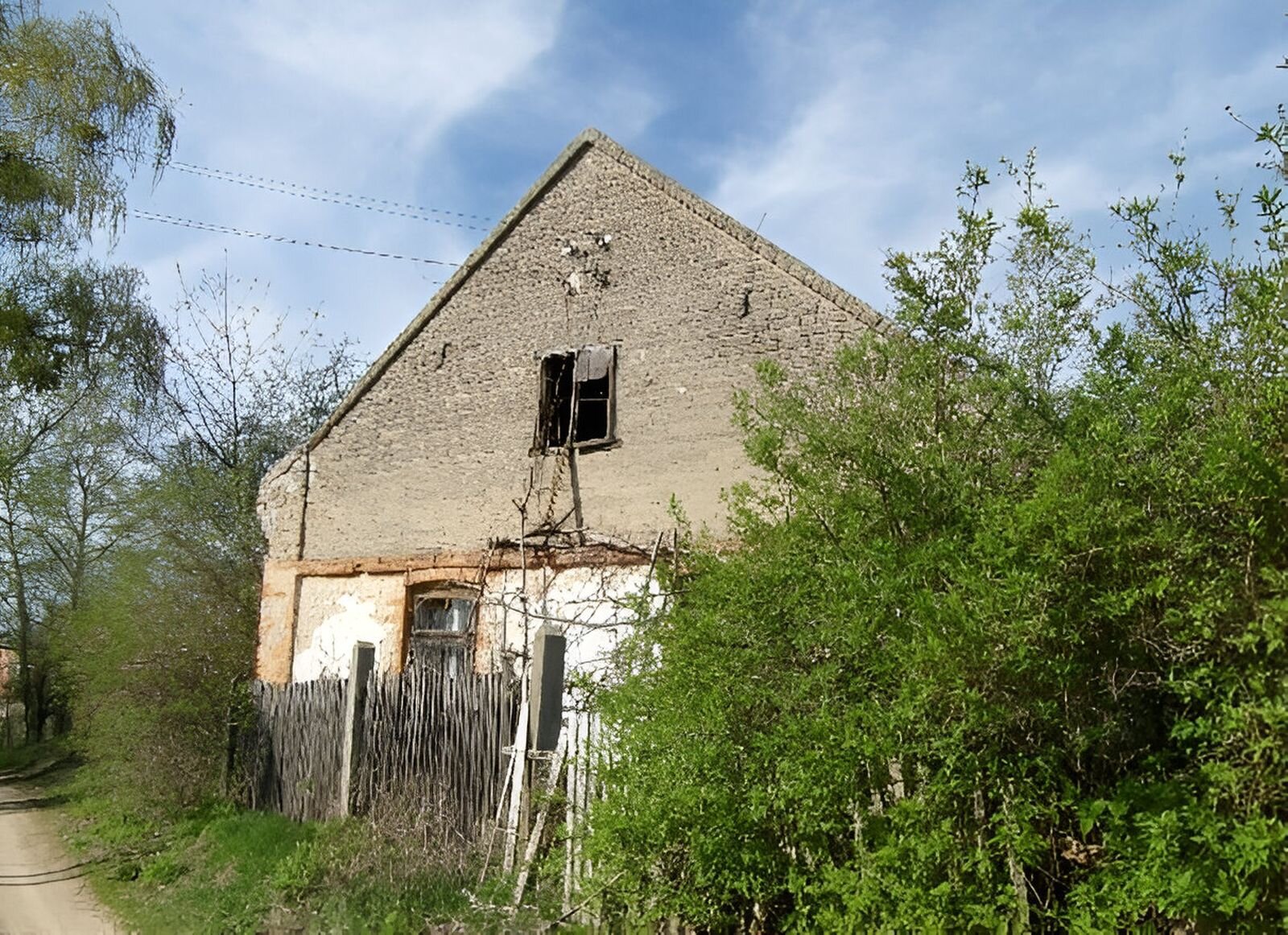 Fotografia przedstawia stary, opuszczony, murowany dom, ogrodzony drewnianym płotem, obok kępiaste krzewy. W domu okna są powybijane. 