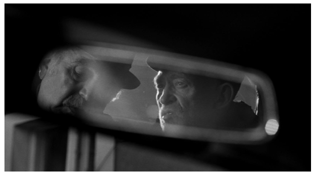 Zdjęcie przedstawia wnętrze taksówki i przednie lusterko. Widać w nim twarze dwóch rozmawiających mężczyzn.