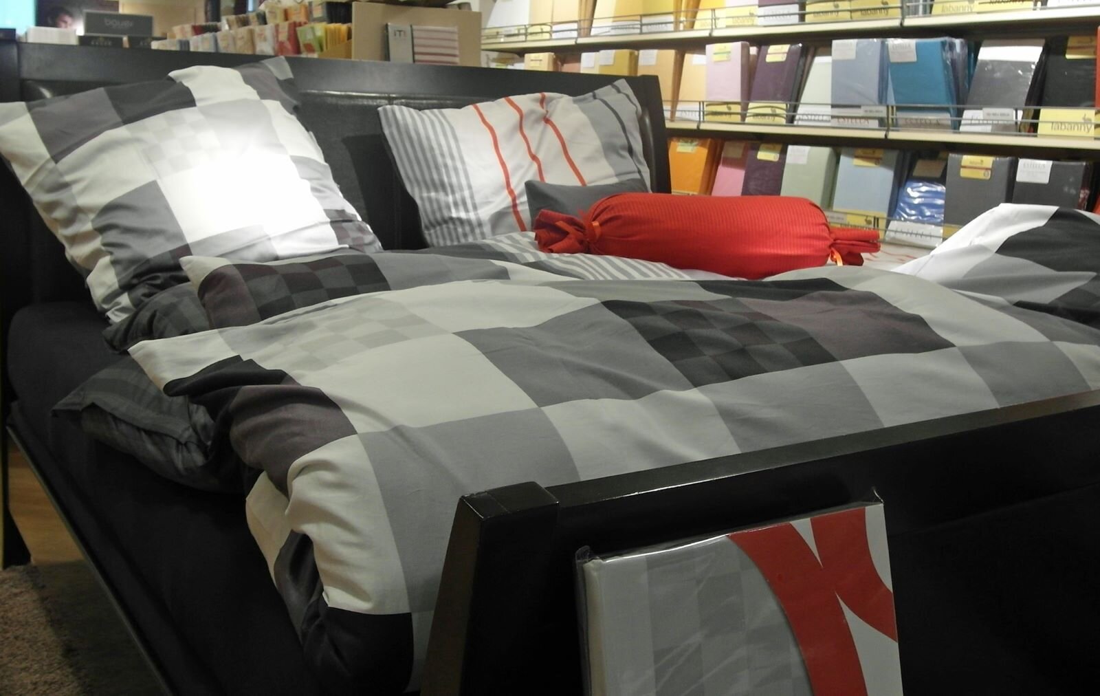 Na zdjęciu przedstawiona jest pościel, leżąca na łóżku. Ma ona wzór w dużą biało‑szaro‑czarną kratę Łóżko stoi w pokoju, w salonie sklepowym. W tle znajdują się półki z kolorowymi, zapakowanymi kompletami pościeli.