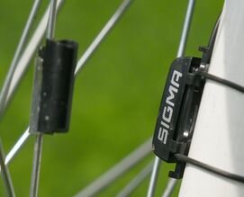 Ilustracja przedstawia dwa czujniki, które są częścią licznika rowerowego.  Jeden z nich zamontowany na szprysze rowerowej, a drugi na widelcu przedniego koła. Elementy zamontowane są na tej samej wysokości. Kiedy elementy mijają się, wytwarzany jest impuls przekazywany do głównej części licznika rowerowego, na którym pokazywane są parametry ruchu. 