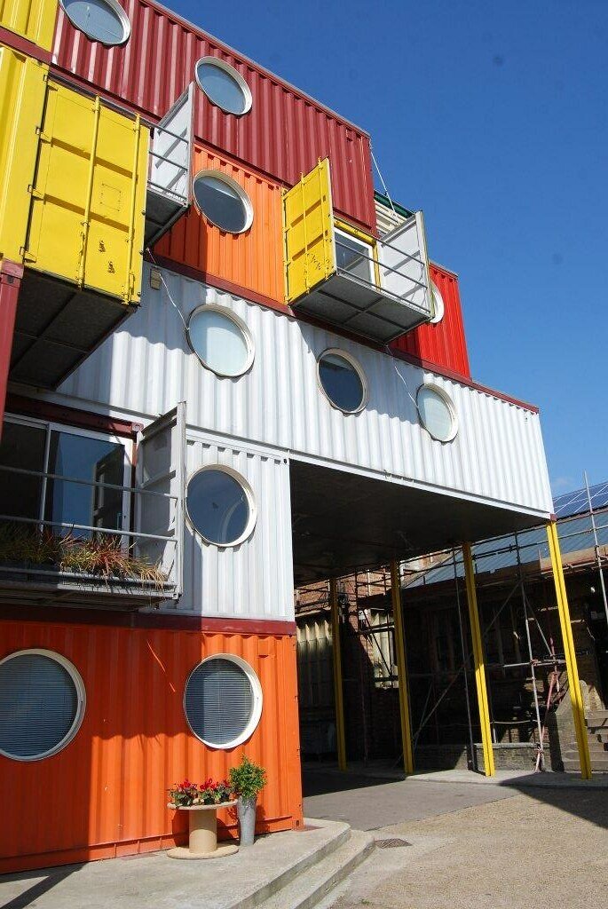 Fotografia przedstawia dom zbudowany z metalowych kontenerów, połączonych ze sobą i tworzących 5‑poziomową budowlę. Część dolnej kondygnacji jest wsparta na żelaznych słupach, tworząc podcień. Każdy kontener ma ściany z blachy falistej i okrągłe okna. W niektórych miejscach zainstalowano małe balkoniki z metalowych ścianek. Przed domem są betonowe schodki, na stojaku przy ścianie domu ustawiono kwiaty.