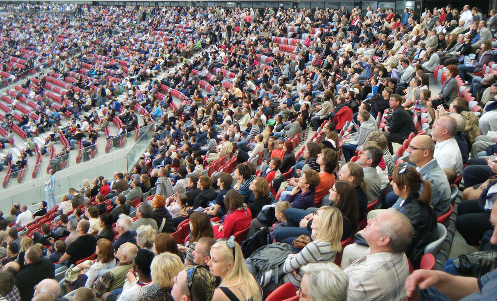 Na zdjęciu ludzie w różnym wieku siedzący na widowni dużej hali lub stadionu.