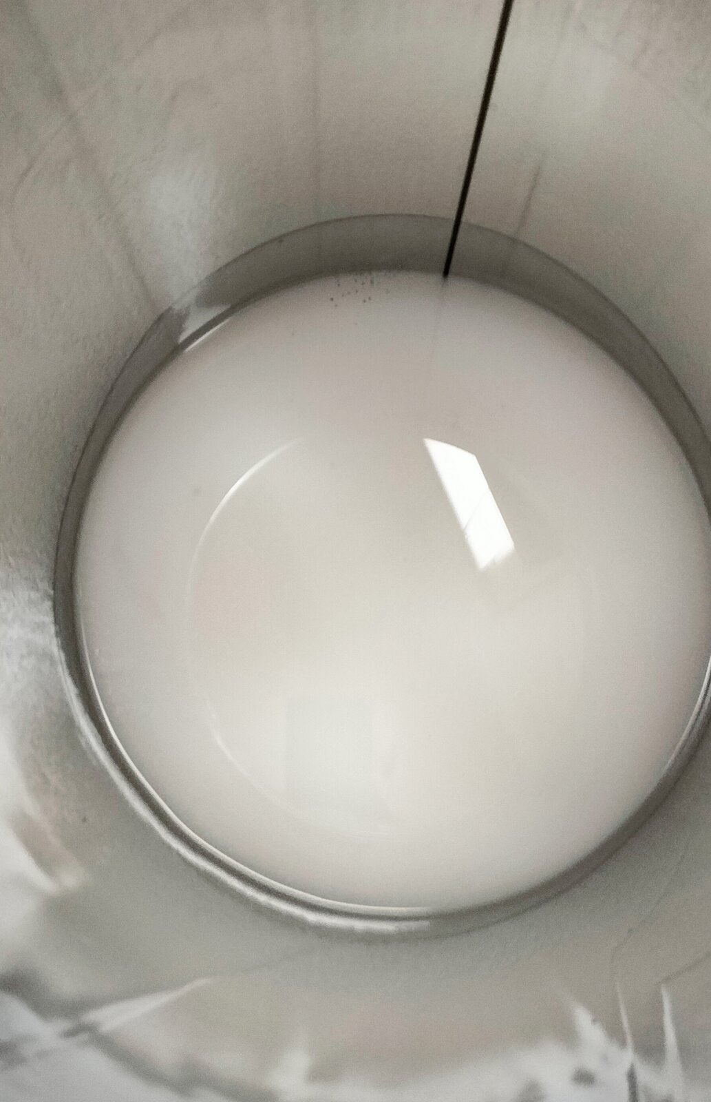 Zdjęcie przedstawia fragment cylindrycznego pojemnika z lakierem do drewna. Widoczny jest z góry srebrny pojemnik, w środku którego znajduje się biały, błyszczący płyn.