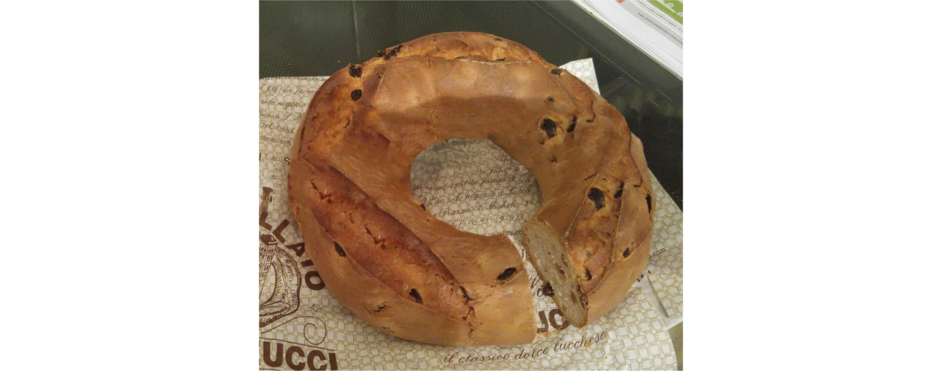 Zdjęcie przedstawia popularne ciastko z Lukki. Ciasto jest koloru brązowego ciemnymi kropkami ma kształt owalny z dużą dziurką w środku. Ciasto jest napoczęta.