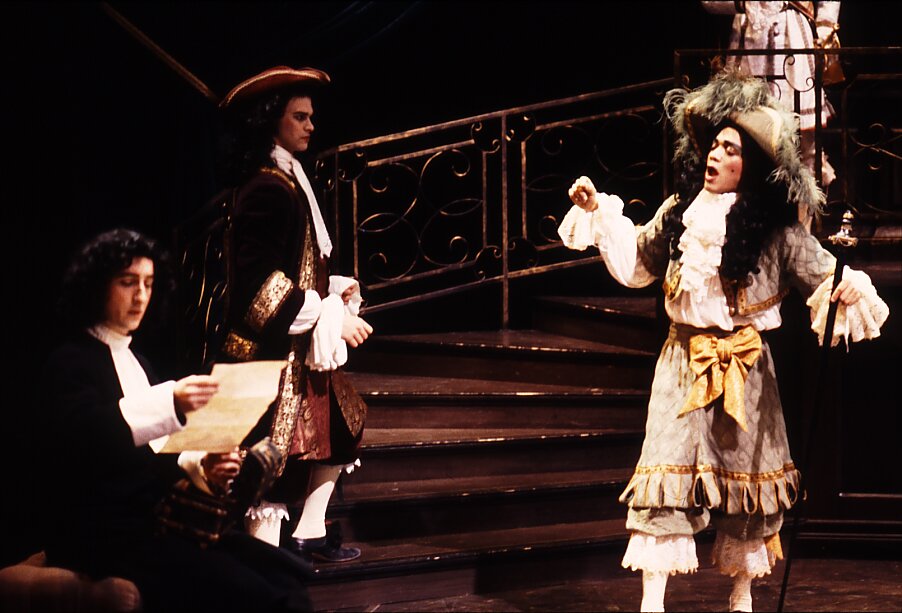 Fotografia przedstawia trzech mężczyzn stojących na scenie. Postać ukazana po lewej stronie czyta list. Mężczyzna ukazuje prawy profil owalnej twarzy. Drugi mężczyzna ukazuje lewy profil. Postać śpiewa. Mężczyzna ma na głowie kapelusz z piórem. Jest ubrany w koszulę z żabotem i spodnie obszyte koronką. Trzeci mężczyzna opiera plecy o barierkę schodów. Ukazuje prawy profil, patrząc na śpiewającego.