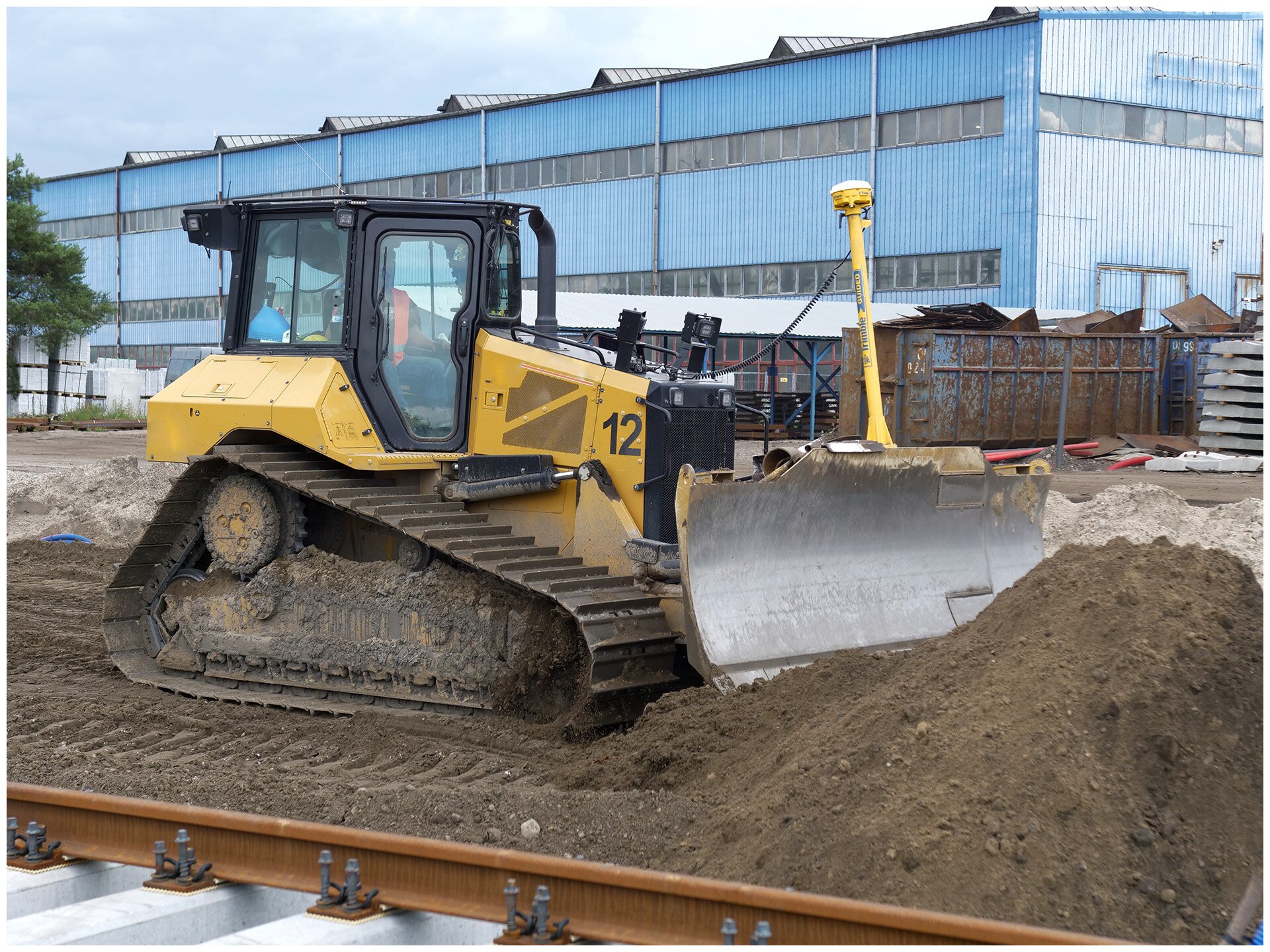 Ilustracja przedstawia spycharkę. Osadzona jest na podwoziu gąsiennicowym, a na przodzie pojazdu jest zamontowany spych. Na zdjęciu widzimy jak spycharka spycha piach na budowie.