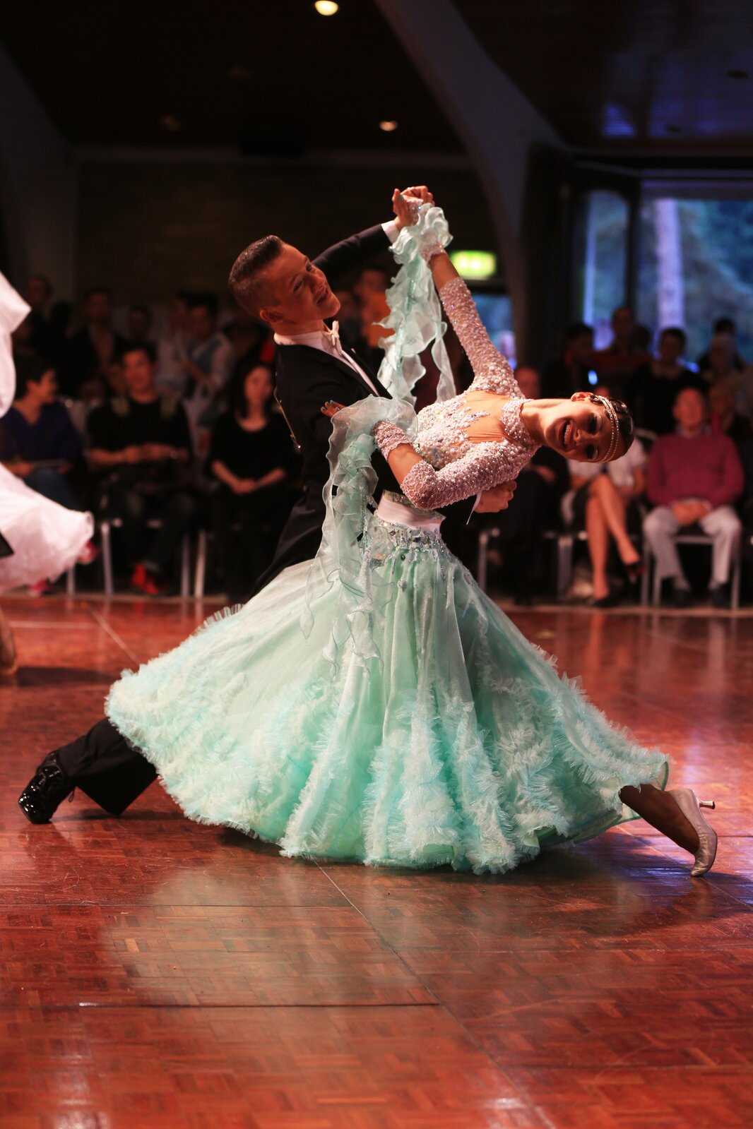 Fotografia przedstawia parę w tańcu. Kobieta ma długą, rozkloszowaną suknię. Góra sukni jest z białej koronki wyszywanej białymi koralikami, w pasie jest biała wstążka, a dół miętowy z falbankami. Na nadgarstkach ma opaski z materiału jak góra sukni, do którego są przyszyte długie kawałki miętowego materiału. Jej paznokcie są pomalowane na kolor miętowy. Na stopach ma srebrne, zakryte buty na niskim obcasie. Jej włosy są ciemne, a na czole ma podwójną, białą opaskę. Mężczyzna ma czarną marynarkę, spodnie, zakryte, wiązane buty oraz białą koszulę i muszkę. Ma krótkie, ciemne włosy. Mężczyzna trzyma lewą dłonią prawą dłoń kobiety, a prawą dłoń ma na jej lewej łopatce. Kobieta lewą dłoń trzyma na prawym ramieniu mężczyzny i odchyla się do tyłu. Oboje mają nogi w dużym rozkroku. Parkiet jest ciemnobrązowy. W tle na krzesłach siedzi wiele osób. Tło jest ciemne, niewyraźne. 