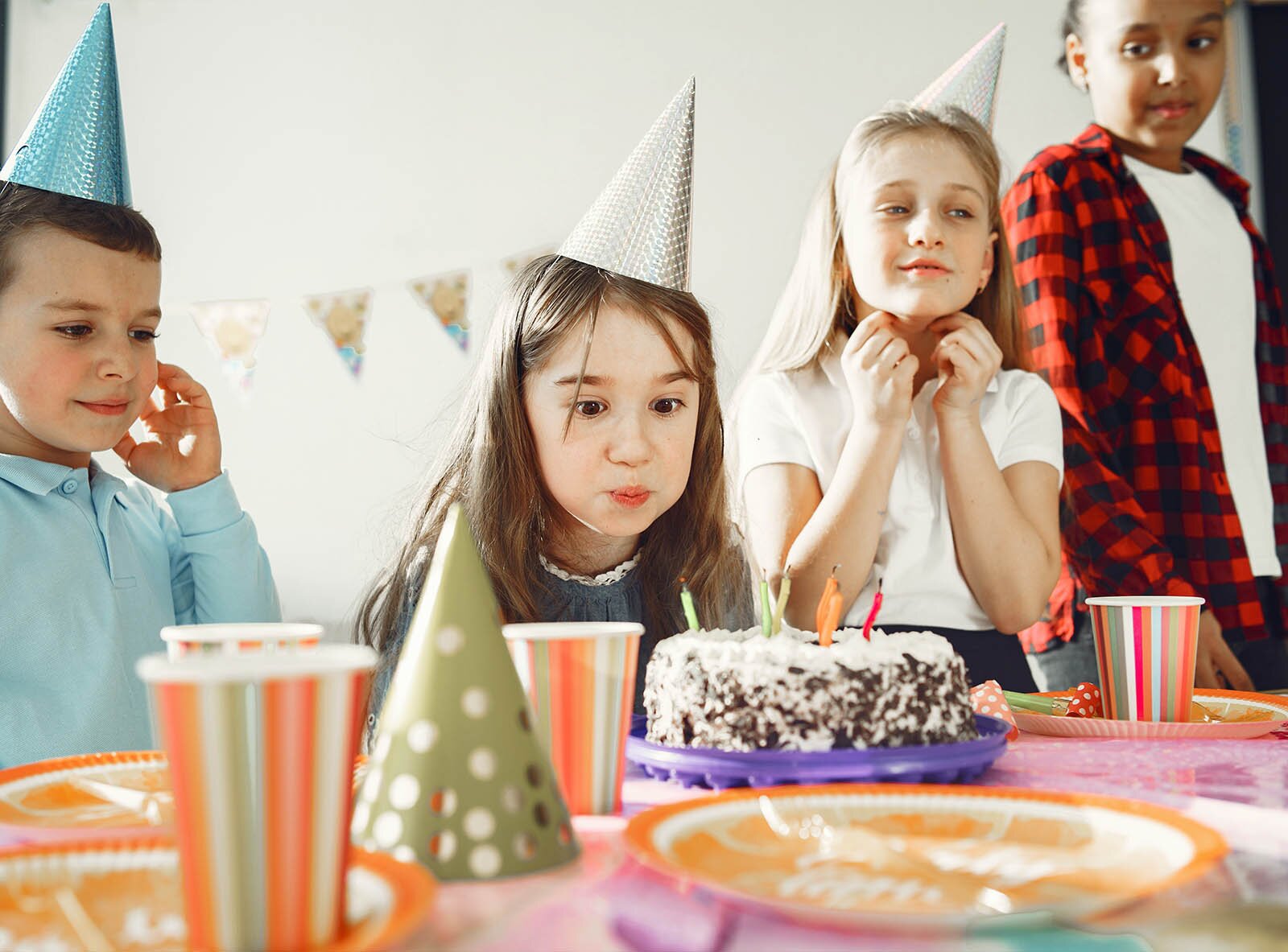 Zdjęcie przedstawia przyjęcie urodzinowe. Widoczny jest tort ze świeczkami, które zdmuchuje dziewczynka w szkolnym wieku. Obok stołu widoczni są goście solenizantki w tym samym wieku. Wszyscy mają trójkątne czapeczki na głowach. 