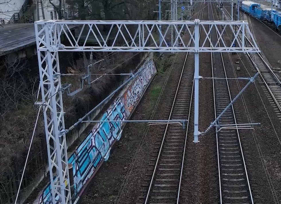 Zdjęcie przedstawia metalową konstrukcję wsporczą. Jest to stalowy słup z kratownicą stojący obok toru. Na szczycie słupa znajduje się poziome ramię, pod którym zawieszone są liny nośne i przewody jezdne.