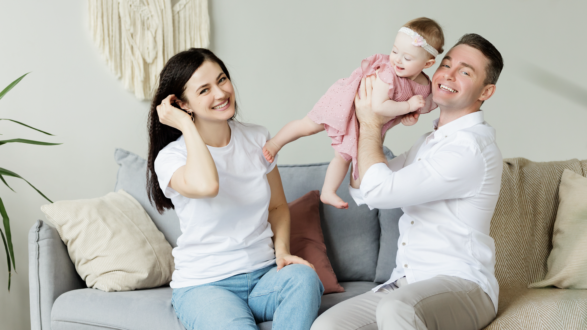 Zdjęcie przedstawia pokój dzienny. W centralnym miejscu stoi sofa, na której leżą poduszki i koc. Na sofie siedzi rozbawiona rodzina. Po lewej stronie śmiejąca się kobieta. Po prawej stronie mężczyzna podrzucający do góry niemowlę ubrane w sukienkę.