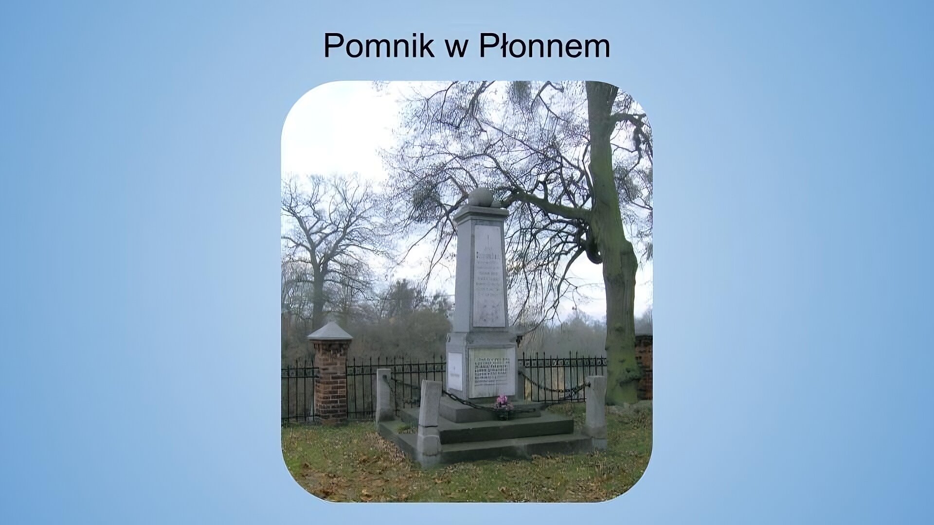 Jasnoniebieskie tło. Na górze napis: pomnik w Płonnem. Poniżej fotografia przedstawiająca pomnik w kształcie wysokiej, wąskiej bryły zwężającej się ku górze. Pomnik ma szary kolor, a przyczepione do niego tablice są jaśniejsze od reszty bryły. U podstawy obiektu znajdują się trzy schodki, na najwyższym z nich leżą różowe kwiaty. Wokół schodków wkopane są cztery niskie słupki, do których przyczepione są łańcuchy tworzące ogrodzenie. Wokół pomnika widoczna jest przystrzyżona trawa i opadłe liście. W tle widać metalowy płot z ceglanymi słupkami oraz drzewa.