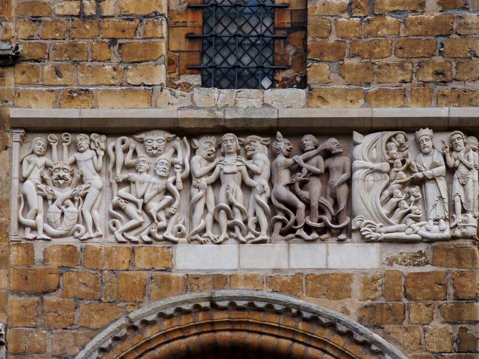 Romański fryz kamienny na elewacji katedry: ciąg płaskorzeźb z gęsto ułożonymi postaciami ludzkimi i fantastycznymi stworami, ukazujący sceny moralizujące z potępionymi i zbawionymi; jasny kamień kontrastuje z surowym murem z bloków.