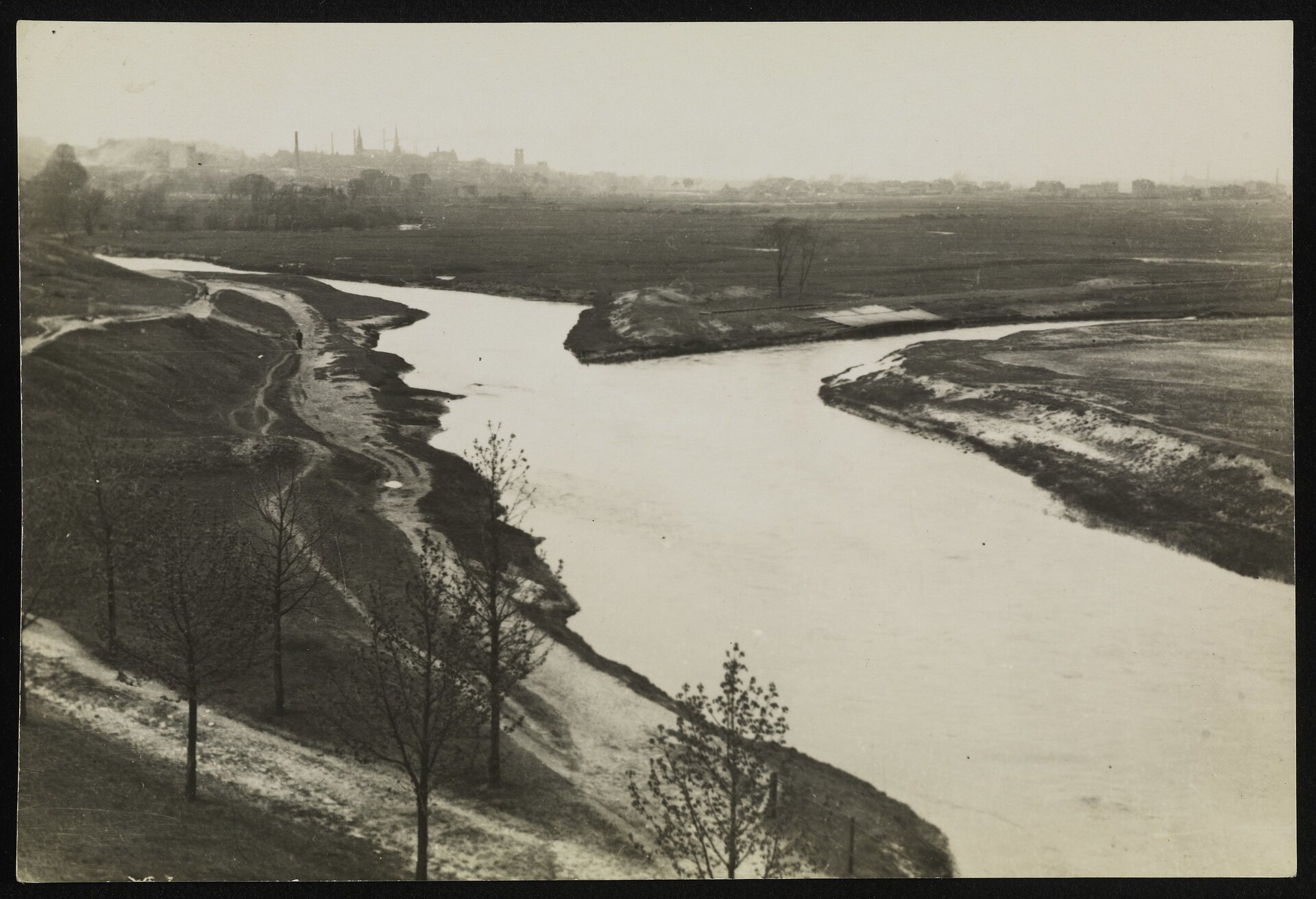 Czarno‑białe zdjęcie przedstawiające dopływ Białej i Czarnej Przemszy do Przemszy. Od lewego górnego rogu do prawego dolnego rogu fotografii biegnie szersza rzeka. W połowie wpływa do niej węższa rzeka, która biegnie od prawego górnego rogu na skos, w dół. Brzegi obu rzek są podmokłe. Na pierwszym planie, po lewej stronie zdjęcia widać nieutwardzoną drogę z drzewami po bokach. Po prawej stronie zdjęcia, za rzeką, widać pola. W tle (na wprost) widoczne są zabudowania miasta. 