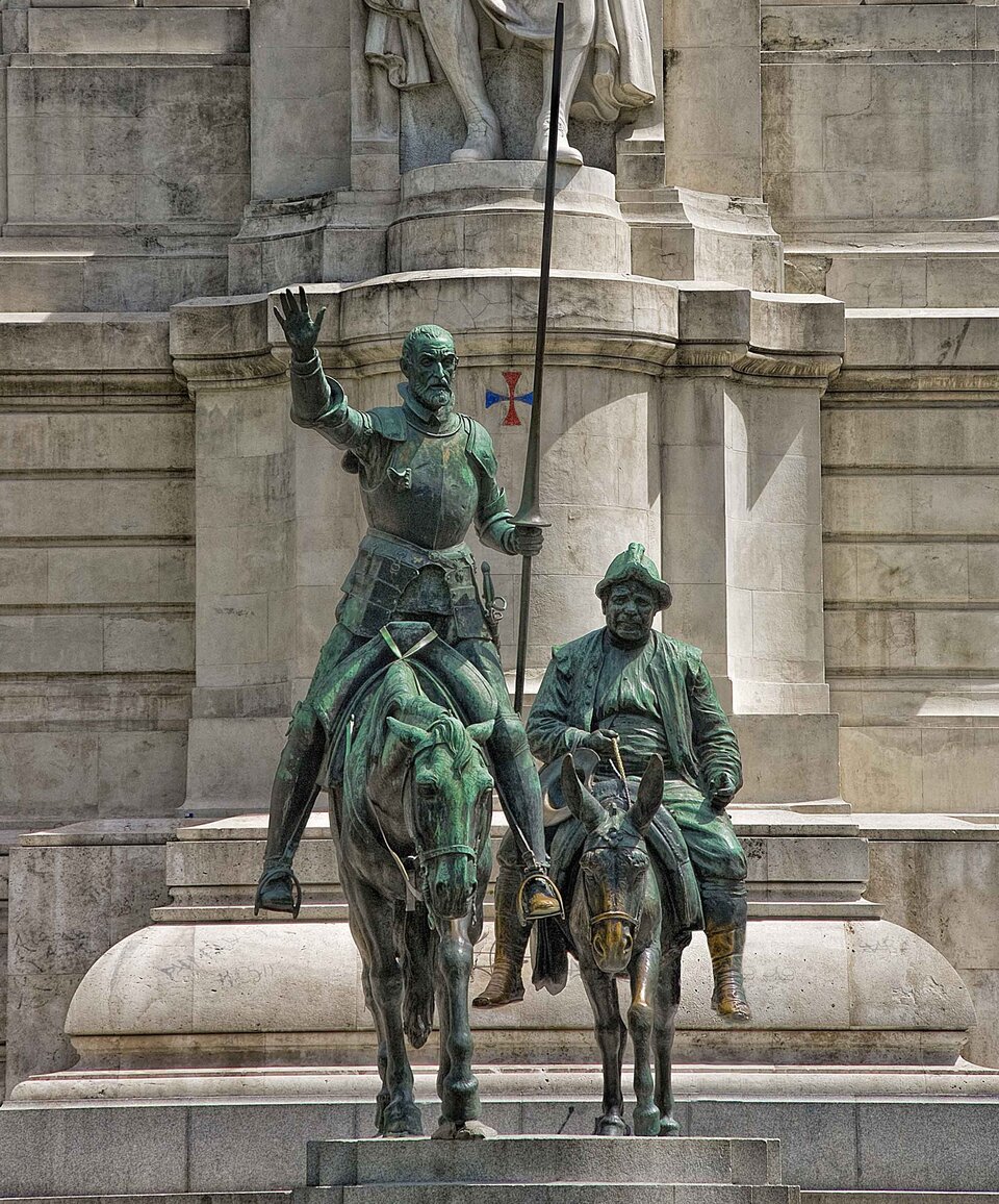 Na zdjęciu znajduje się pomnik Don Kichota i Sancho Pansy. Po lewej  stronie siedzi na koniu rycerz z krótkimi włosami i grzywką opadającą na czoło oraz z brodą. Jest ubrany w zbroję. Przy boku ma szablę. W prawej dłoni trzyma lancę. Lewą rękę unosi do góry. Po jego lewej stronie siedzi na ośle niski i pulchny giermek. Na głowie ma kapelusz. Jest ubrany w koszulę przepasaną pasem, spodnie, kontusz i buty do połowy łydki. W prawej dłoni trzyma lejce. Postacie pomnika umieszczone są na cokole. W tle widać kamienną ścianę.