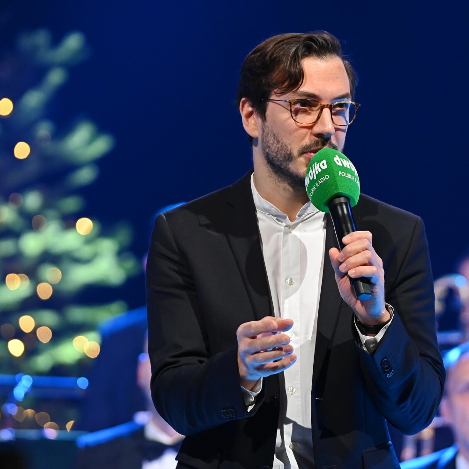 Ilustracja przedstawia fotografię Andrzeja Zielińskiego. Mężczyzna trzyma w lewej ręce zielony mikrofon z białym napisem „Dwójka‑Polskie Radio”. Przemawiając do mikrofonu, gestykuluje prawą ręką.  Ubrany jest w czarną, klasyczną marynarkę, białą koszulę oraz korekcyjne okulary. Zamyślony mężczyzna ma czarne włosy z grzywką opadająca na prawy bok i delikatny zarost. W tle znajduje się siedząca publiczność, a za nimi po lewej stronie sporych rozmiarów, zielona choinka z ciepłym oświetleniem. Tło za nim jest rozmazane w granatowym kolorze.