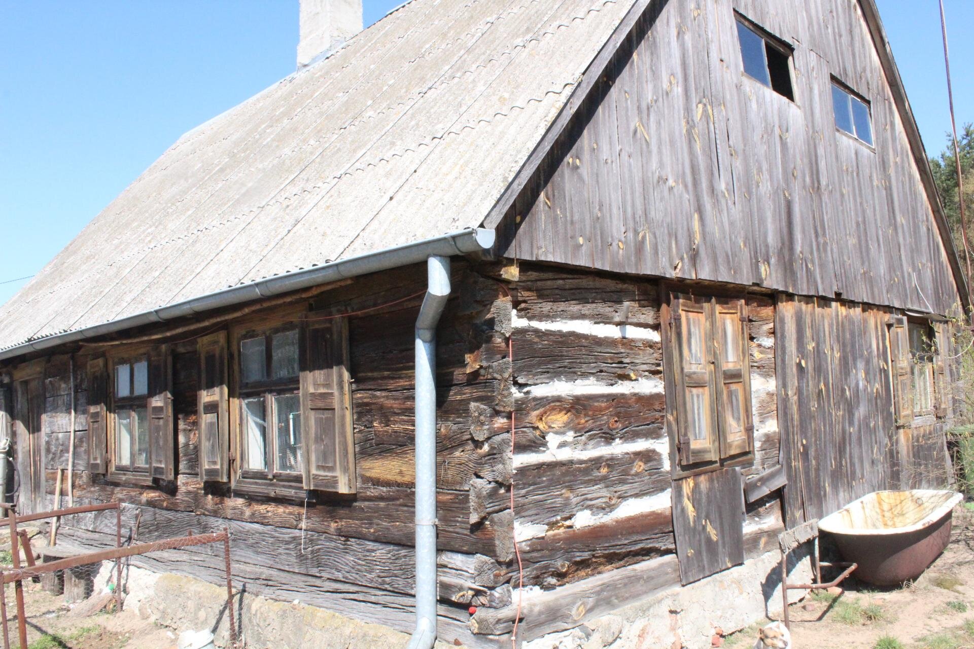 Zdjęcie przedstawia drewnianą, piętrową chatę ze skośnym dachem. Ma ona po jednej stronie dwa okna z drewnianymi okiennicami po bokach, a z drugiej strony okno i drewniane, podwójne drzwi. Na piętrze znajdują się dwa małe okna. Z prawej strony domu stoi zardzewiała, biało‑brązowa wanna. Niebo jest błękitne, bezchmurne. Zdjęcie zostało wykonane w świetle dziennym.