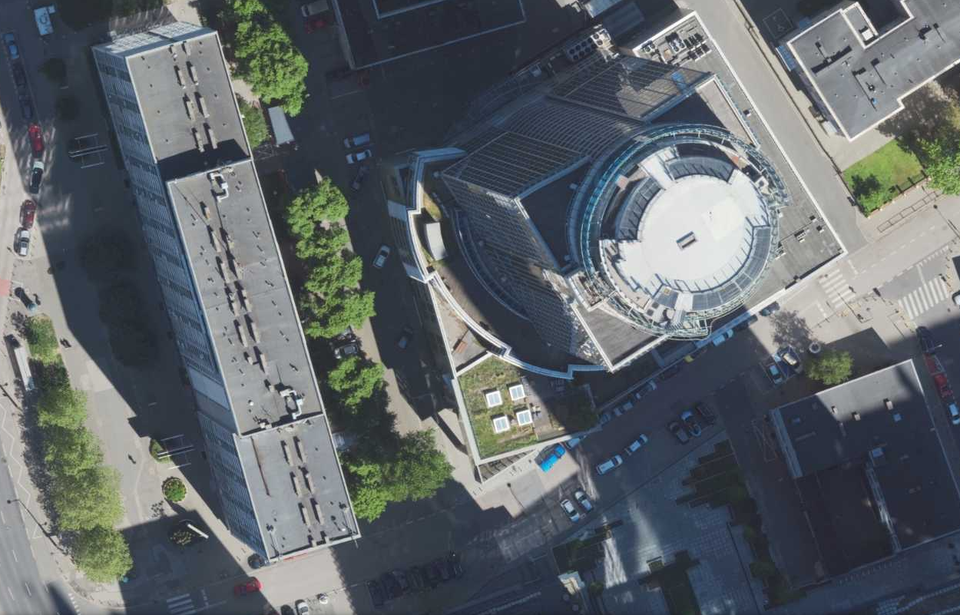 Ilustracja przedstawia zdjęcie z lotu ptaka na helipad znajdujący się na wieżowcu Spectrum Tower, w centrum Warszawy. Wokół niego są niższe budynki. Widoczne są liczne miejsca parkingowe.