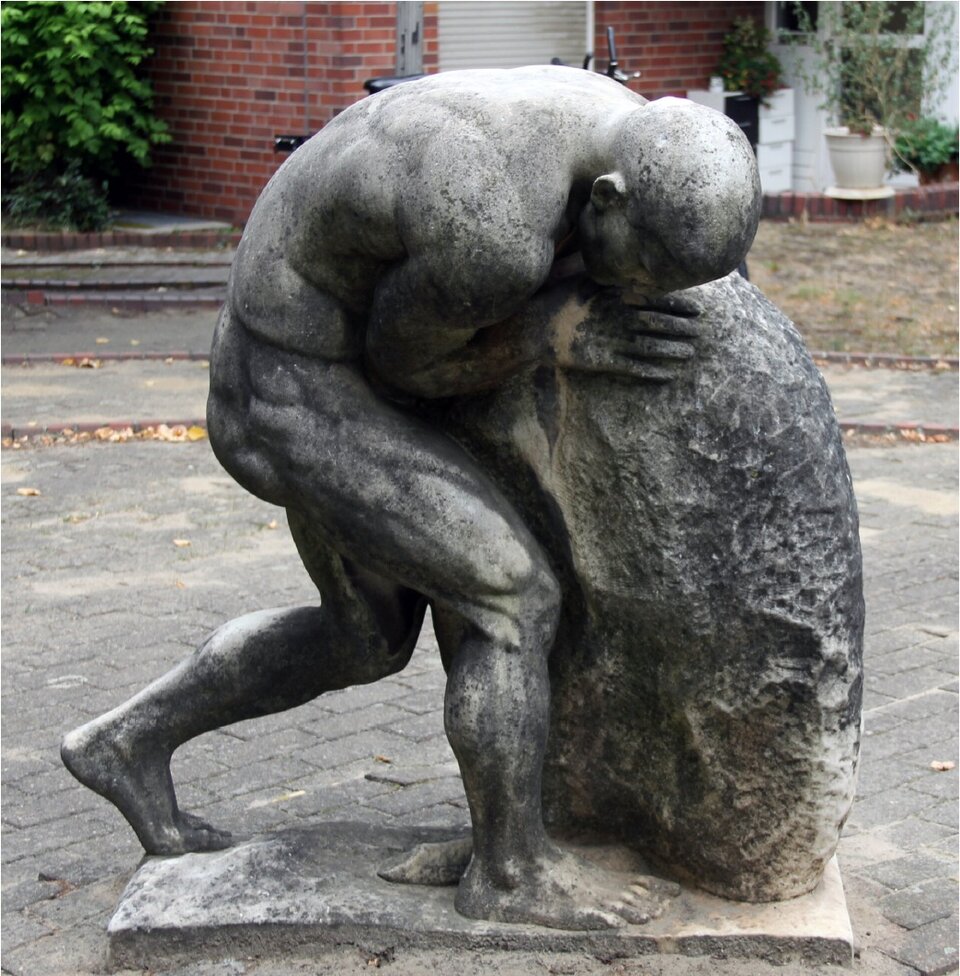 Zdjęcie przedstawia rzeźbę pochylonego, muskularnego mężczyzny, który próbuje pchać przed sobą duży głaz.