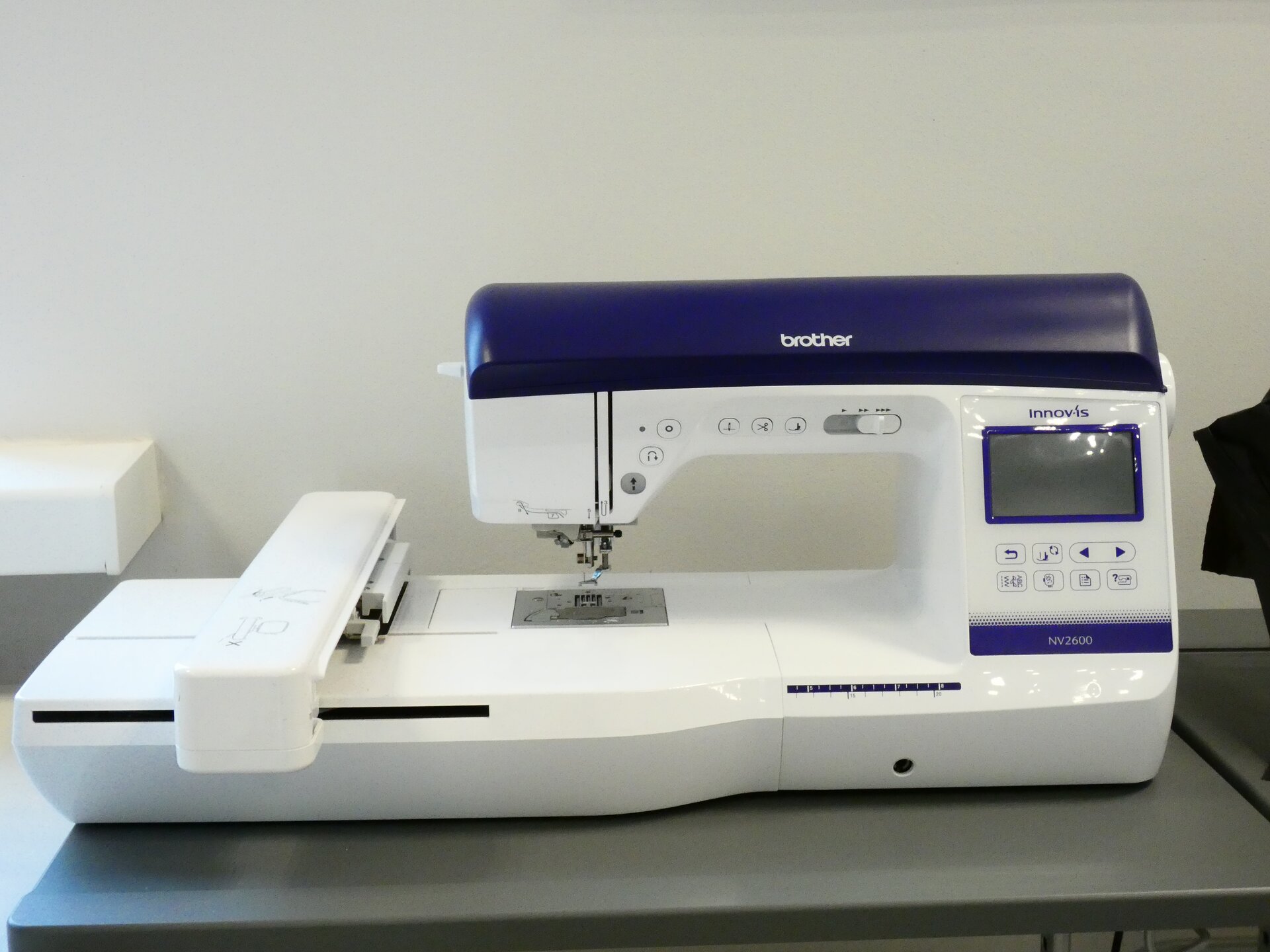 Zdjęcie przedstawia białą maszynę do szycia. Jej góra jest granatowa. Po prawej stronie znajduje się panel sterujący z ekranem, a po lewej podłużna, ruchoma część położona w poprzek maszyny.