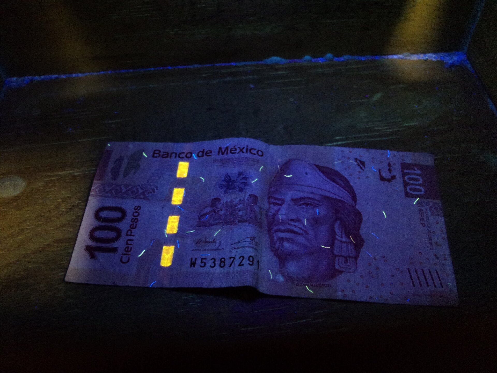 Zdjęcie przedstawia banknot sto meksykańskich pesos oświetlony lampą kwarcową. Wyraźnie widoczne są cztery prostokąty narysowane w linii w poprzek banknotu, oraz chaotycznie zaznaczone krótkie kreski, podczas gdy reszta banknotu jest zacieniona, tak jak i jego otoczenie.