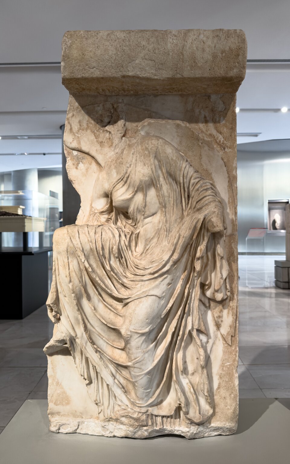 Ilustracja przedstawia płaskorzeźbę „Nike zawiązująca sandał” . Na zdjęciu widoczny jest fragment reliefu z wyrzeźbioną w technice mokrych szat Nike. Postać kobiety ukazana jest w trakcie zdejmowania sandała. Zgięte, prawe kolano unosi się do góry, a prawa ręka sięga ku stopie. Cała sylwetka pochyla się ku przodowi. Płaskorzeźba nie jest kompletna, nie zachowały się głowa i fragment lewej ręki z dłonią.