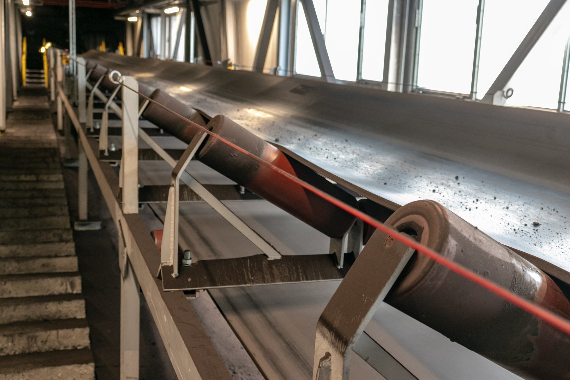 Na zdjęciu widać fragment przenośnika taśmowego czyli urządzenia wykorzystywanego do transportu materiału na dłuższe odległości z podstawowymi jego podzespołami czyli krążnikami, taśmą oraz kozłem górnym. Krążniki są to obrotowe wałki, po których przesuwa się taśma przenośnika.Przenośnik taśmowy jest to podłużne urządzenie mechaniczne na górze którego znajduje się czarna taśma. Podstawą przenośnika taśmowego jest stalowa konstrukcja kratowa czyli trasa. Konstrukcja ta zbudowana jest powtarzalnych członów modułów których podstawą są ceowniki. Ceownik jest to wyrób hutniczy, walcowany lub gięty z metalu. Wyroby te są w postaci prostych odcinków. Przekrój poprzeczny czyli profil ceownika przypomina literę C.Z boku konstrukcji czyli na boku trasy znajdują się metalowe elementy w kształcie ceowników, które biegną pionowo w górę. Końcówki tych elementów są zakrzywione w kierunku taśmy czyli w kierunku środka przenośnika. Są to kozły górne. Kozły górne są rozmieszczone po oby stronach trasy. Na koźle górnym widać krążnik górny. Krążnik jest w kształcie czerwonego walca. Na krążnikach znajduje się długa taśma która biegnie przez całą długość przenośnika.Od boku trasy odchodzą równomiernie stalowe elementy w kształcie odwróconej litery L. Przedstawiony na zdjęciu przenośnik znajduje się we wnętrzu obiektu budowlanego. Po lewej stronie zdjęcia znajduje się przejście na końcu którego widać schody z żółta poręczą. Po prawej stronie zdjęcia widać okna.