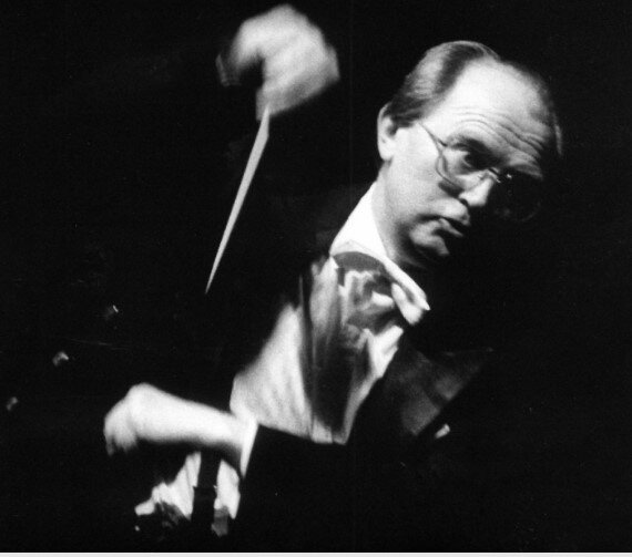 Ilustracja przedstawia dyrygenta, mężczyzna ubrany w czarny frak i białą koszulę na ciemnym tle.