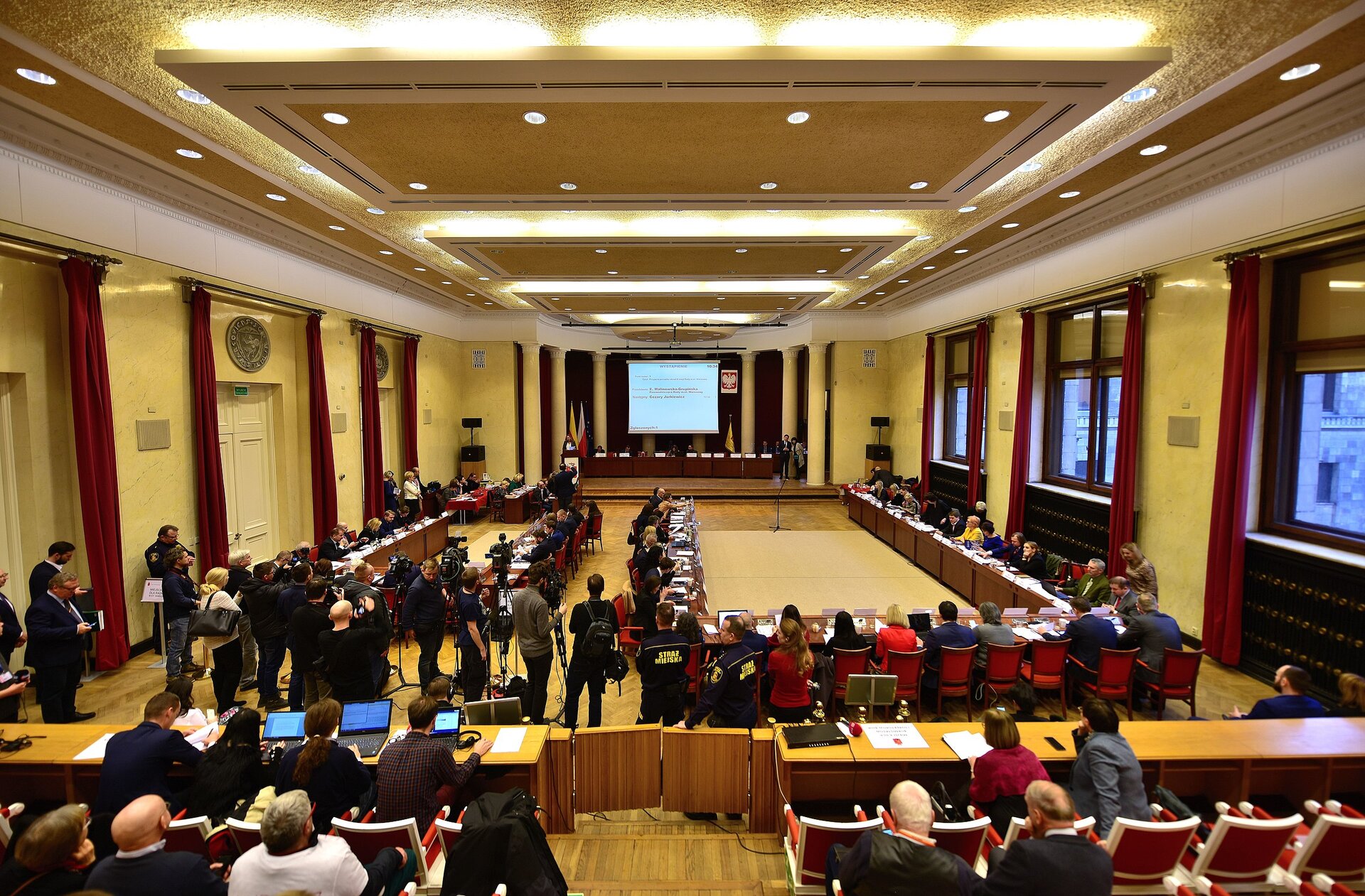 Zdjęcie przedstawia salę, w której odbywa się sesja rady miasta stołecznego Warszawy. Sala jest przestronna. Na suficie jest dużo punktów świetlnych. Na centralnej ścianie stoi długi stół, przy którym siedzą osoby. Za stołem jest ekran projektora. Obok niego wisi godło Polski. Wokół pomieszczenia biegną stoły, za którymi siedzą radni. Przy stołach stoją z kamerami przedstawiciele mediów. Widoczna też jest publiczność, która siedzi na wprost stołu. 