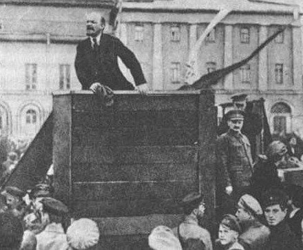 Na zdjęciu stoi drewniana mównica. Za nią stoi Włodzimierz Lenin i przemawia do zebranych ludzi. W oddali widać budynek.