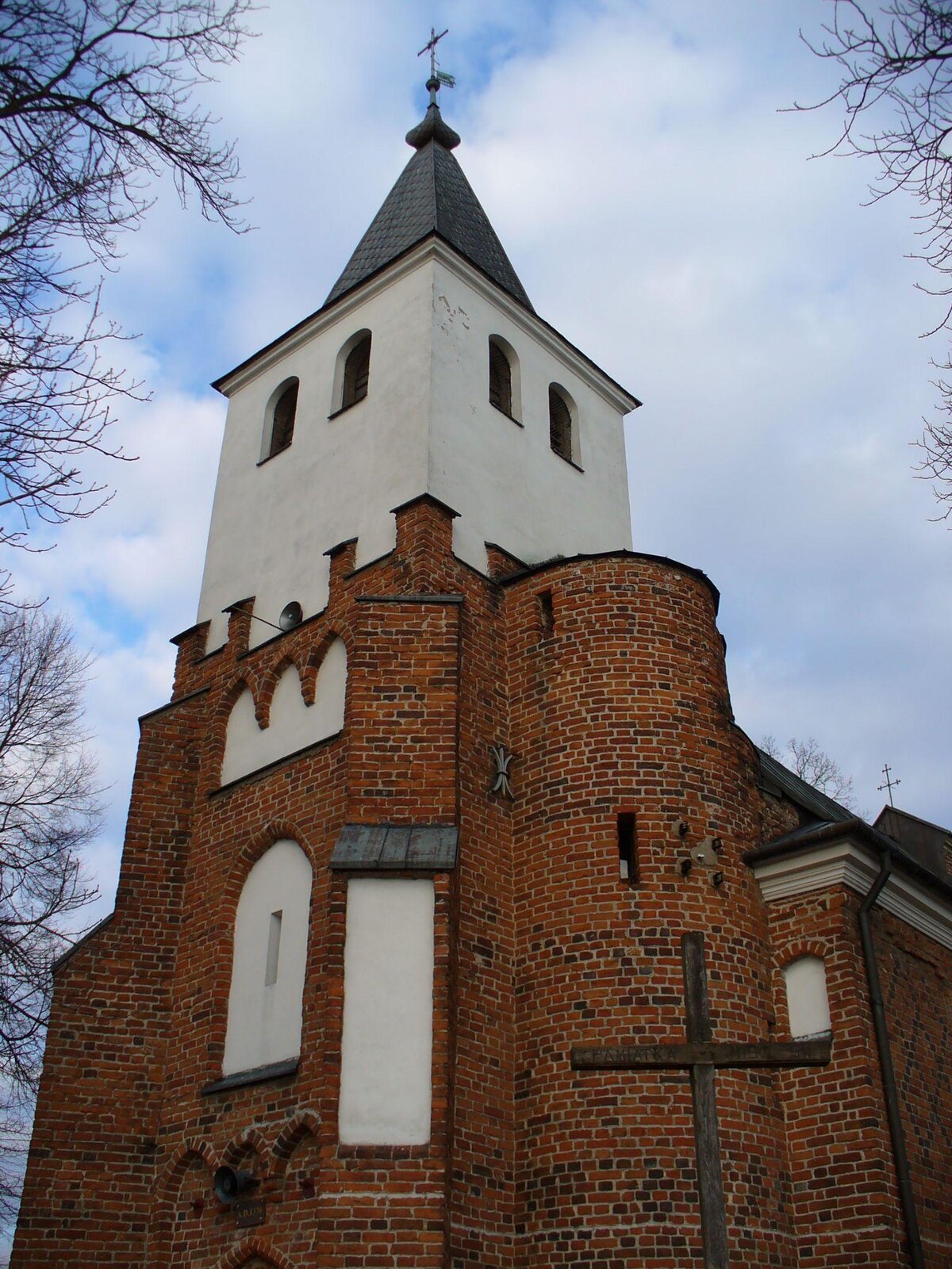 Zdjęcie przedstawia przód kościoła. Na pierwszym planie znajduje się wysoka wieża, częściowo obudowana czerwoną cegłą, a częściowo białym tynkiem. Wieża zakończona jest szarym, spiczastym dachem, na którego czubku znajduje się krzyż. Za wieżą widoczna jest nawa główna, której ściany obłożone zostały czerwoną cegłą. Przed kościołem stoi drewniany krzyż. Wokół kościoła widoczne są bezlistne gałęzie. Niebo jest błękitne, częściowo przysłonięte białymi chmurami. Zdjęcie zostało wykonane w świetle dziennym.