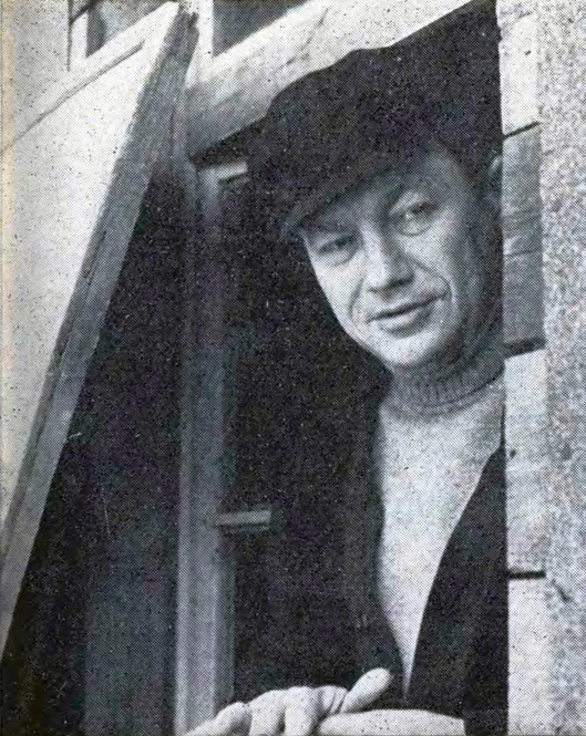 Fotografia przedstawia portret mężczyzny w sile wieku. Postać, wychylając się przez otwarte okno, prezentuje lewy półprofil. Mężczyzna jest ukazany od piersi w górę. Lekko pochyla głowę. Włosy mężczyzny są zasłonięte czapką z krótkim daszkiem. Reżyser ma owalną twarz. Jego czoło jest lekko ocienione, niewysokie. Brwi postaci są cienkie, oczy zmrużone, lekko opuchnięte. Nos mężczyzny jest długi, prosty, dość kanciasty. Usta są szerokie, delikatnie otwarte. Postać ma gładko ogoloną twarz. Jest ubrana w sweter z golfem i marynarkę. Mężczyzna opiera dłonie o parapet okna.