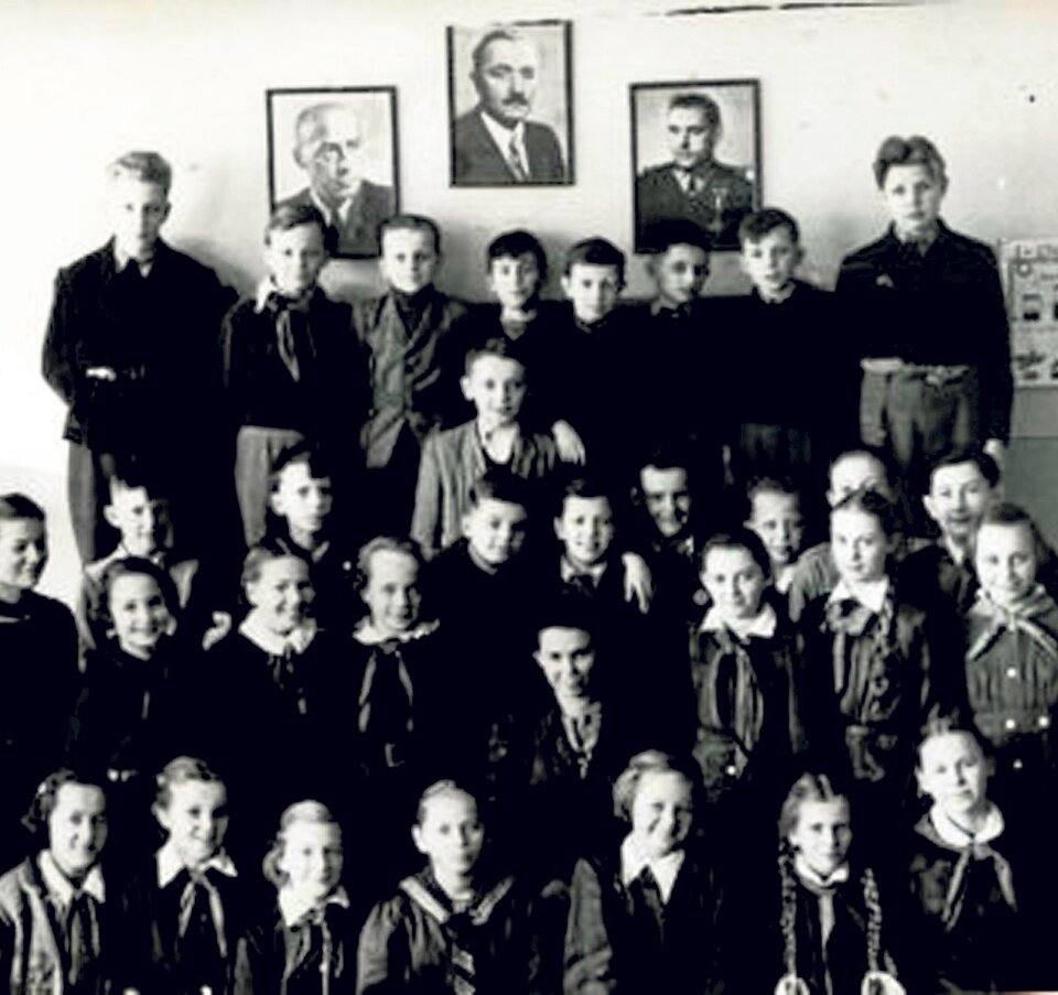 Zdjęcie przedstawia uczniów pozujących do zdjęcia. Część z nich jest w mundurkach szkolnych, część w mundurach harcerskich. Na ścianie, przy której stoją dzieci wiszą trzy portrety mężczyzn będących przywódcami państwowymi i członkami PZPR.