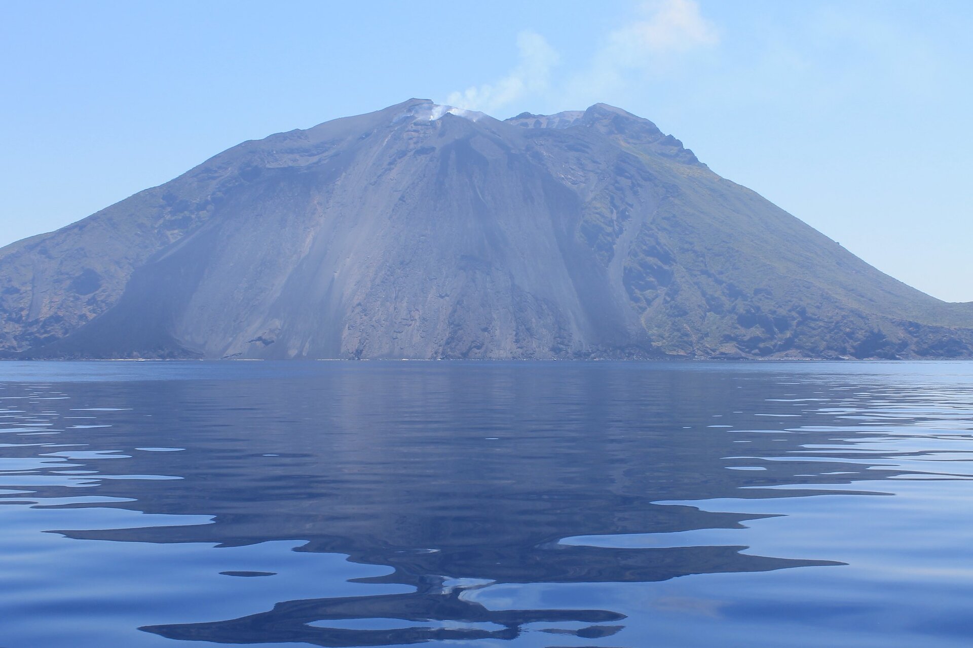 Na zdjęciu jest wulkan położony nad morzem. Na szczycie wulkanu widoczny jest krater, z którego unosi się cienka smuga dymu.  