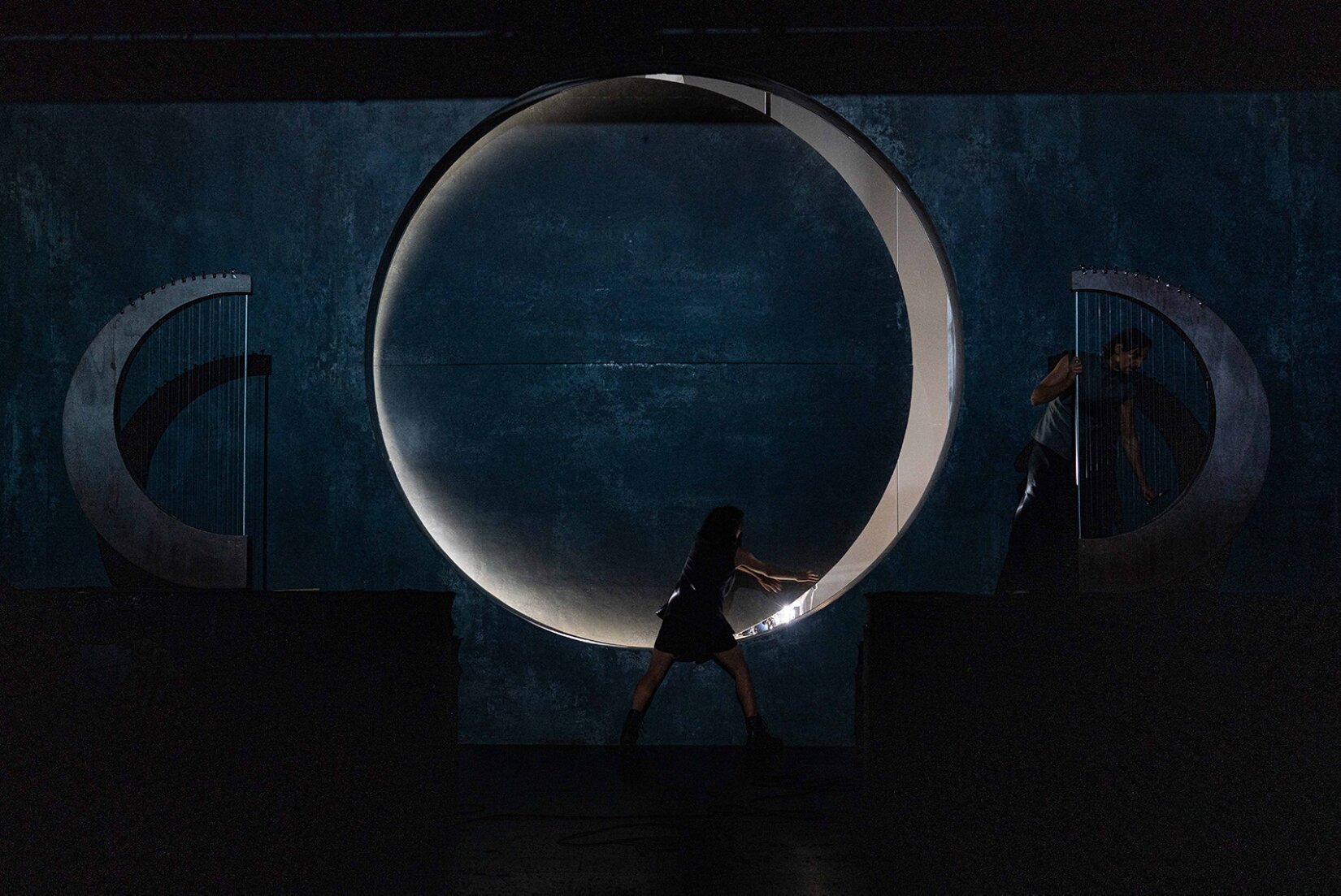 Fotografia przedstawia scenografię do sztuki Elektra. Na scenie znajdują się dwie harfy ustawione po obu jej stronach. W centralnej części wisi sfera, która przepuszcza światło z prawej strony. Scenografia utrzymana jest w ciemnych kolorach. Przy jednej z harf stoi młody mężczyzna, gra na instrumencie. Ubrany jest w długie czarne spodnie oraz szary t‑shirt. Kobieta stoi u dołu sfery i ją odsuwa. 