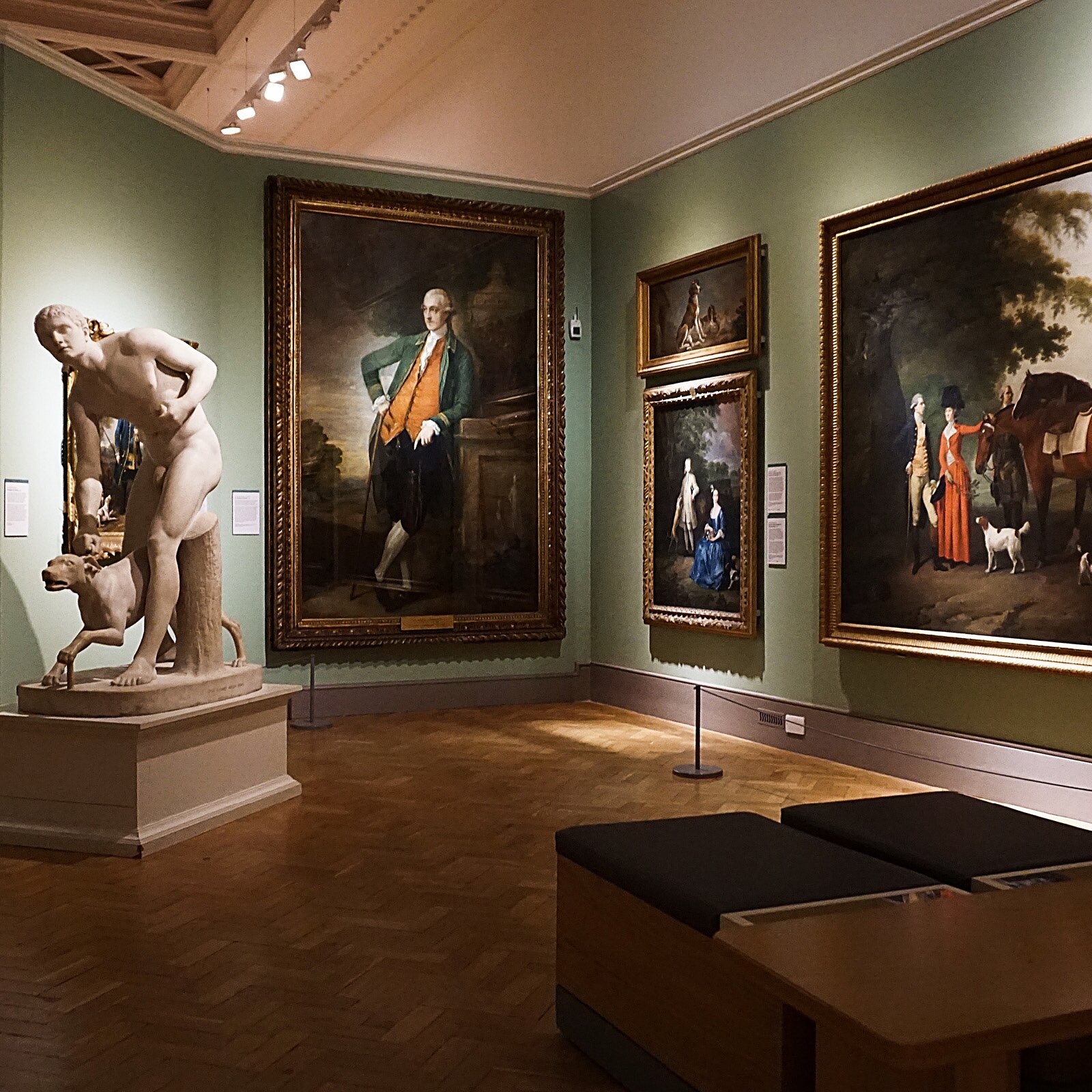 Zdjęcie przedstawia wnętrze muzeum, na którego ścianach wiszą wielkie portrety w złoconych i zdobionych ramach. Widzimy również pomnik nagiego mężczyzny z psem. Na pierwszym planie widać dwa miejsca siedzące z brązowym, miękkim siedziskiem.