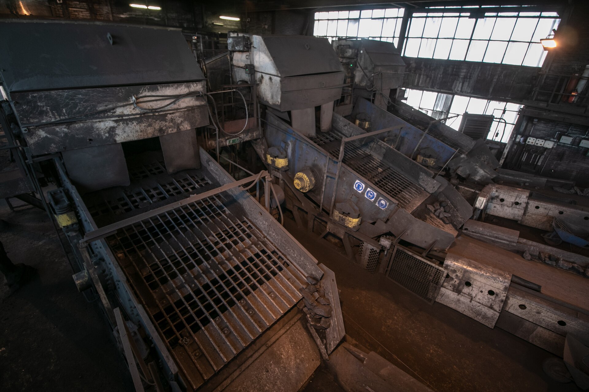 Na zdjęciu widoczne są przesiewacze wibracyjne. Przesiewacz jest to urządzenie służące do rozdzielania na sitach materiału na ustalone grupy wymiarowe ziarn,Na zdjęciu widoczna jest hala w której znajdują się trzy przesiewacze. Przesiewacze stoją obok siebie. Podstawę przenośnika tworzą dwie równoległe usztywnione ściany boczne. Pomiędzy ścianami znajduje się ruszt. Ruszt biegnie pod kątem (jest odchylony od poziomu). Na szczycie rusztu zamontowana jest prostopadłościenna metalowa skrzynia. Po środku skrzyni znajduje się otwór.W tle zdjęcia widoczne sa okna.