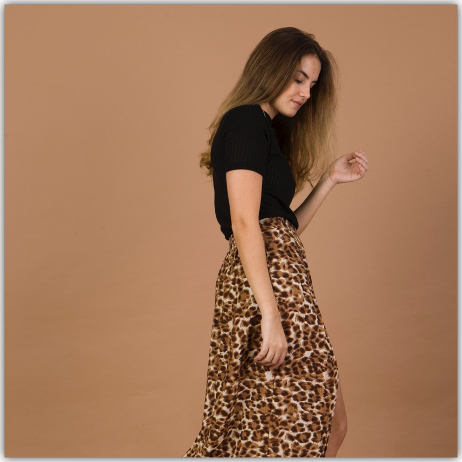 Zdjęcie przedstawia młodą kobietę na jasno brązowym tle. Kobieta ma długie, brązowe włosy. Ma na sobie czarną koszulkę i spódnicę w panterkę. Lewą dłoń skierowaną ma ku górze, patrzy w dół. 