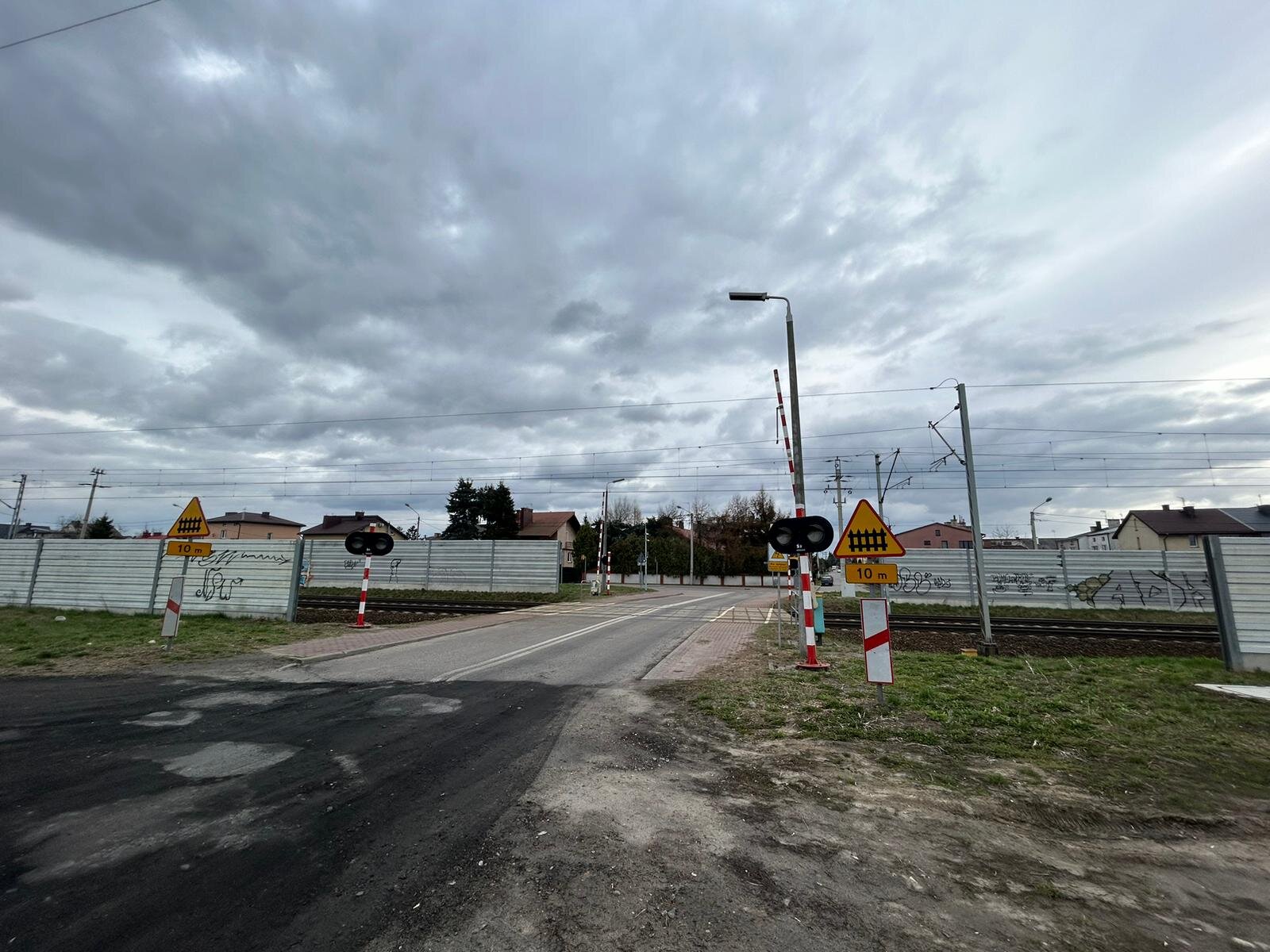 Ilustracja przedstawia przejazd kolejowy. Na ilustracji widoczne są otwarte szlabany rogatek obok nich znajduję się lampa oświetlająca przejazd, Po lewej i prawej stronie znajdują się znaki drogowe. Dwa czerwone białe słupki, na których znajdują się lampki z dwoma światłami. Przed nimi znajduję się znaki informujące o przejeździe kolejowym. Jest to żółty trójkąt z czerwoną obramówką grotem skierowany do góry. Na trójkącie znajduję się grafika płotu zbudowana z pięciu przęseł pionowych, który zakończone są szpicem oraz dwóch poziomych. Pod nim na żółtej tablice znajduję się napis dziesięć em poniżej, biały pionowy znak z czerwoną obramówką w środku znaku poprowadzona jest gruba czerwona linia pod kątem ostrym. Po obu stronach  jezdni znajdują się takie same znaki. Torowisko otacza szary mur.