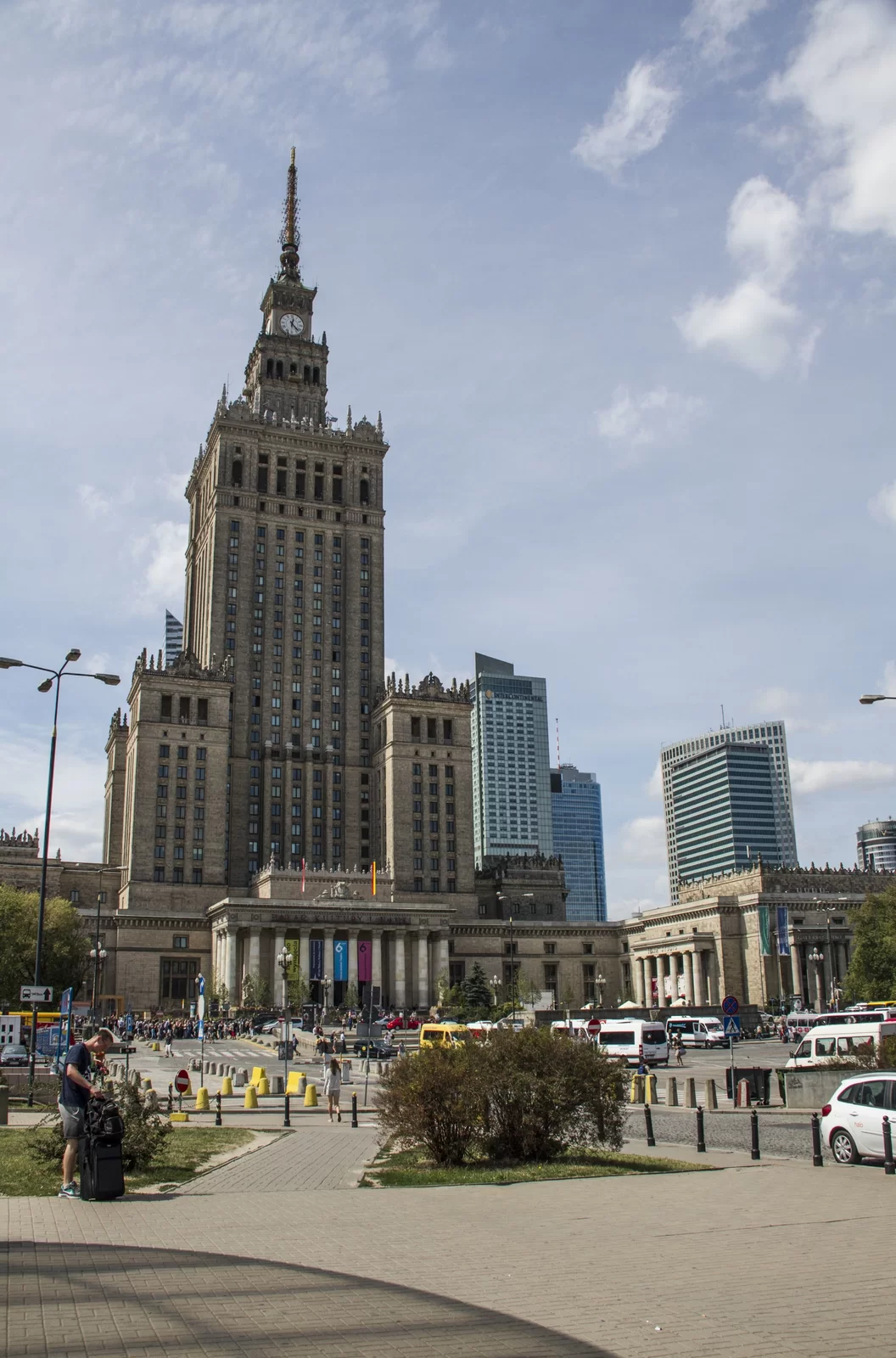 Fotografia przedstawia Pałac Kultury i Nauki w Warszawie. Wysoki budynek w stylu socrealistycznym. Pałac Kultury i Nauki jest najwyższym budynkiem w Polsce, ma 42 piętra o łącznej wysokości ok. 167 metrów. Centralną część stanowi wieża z zegarem, zwieńczona iglicą, której szczyt umiejscowiony jest na wysokości ok. 230 metrów. Wieża posiada cztery narożne wieżyczki przyporowe o wysokości 60 metrów. Połączona jest też z czterema niższymi platformami naziemnymi, przypominającymi staropolski pałac lub dwór. Platformy znajdują się na rogach wieży. Od strony wschodniej, w części środkowej wieży znajduje się także półkolisty ryzalit, w którym mieści się Sala Kongresowa. Długość pałacu wzdłuż ul. Marszałkowskiej wynosi 254 metry, a wzdłuż Alej Jerozolimskich 212 metrów. W pałacu znajduje się łącznie 3288 pomieszczeń o całkowitej powierzchni około 123.000 metrów kwadratowych. Rosyjski architekt, Lew Rudniew, wzorował się na siedmiu moskiewskich drapaczach chmur, dlatego też Pałac Kultury i Nauki architektonicznie jest podobny m.in. do Uniwersytetu Moskiewskiego. Założeniem architekta było, aby architektura zawierała elementy architektury polskiej. Stylizację gmachu oparł na krakowskim renesansie i warszawskim klasycyzmie. Przed wejściem do pałacu stoją dwie rzeźby – pomnik Adama Mickiewicza i Mikołaja Kopernika. Rzeźby znajdują się także wokół pałacu, we wnękach. Główne wejście osłonięte jest kolumnami.