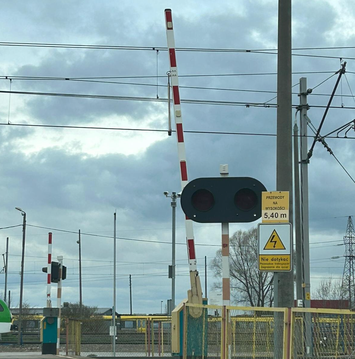 Ilustracja przedstawia rogatkę, rogatka jest koloru czerwono białego i jest otwarta. Pod rogatką znajduję się czerwono biały słup, na którym umieszczono lampę z dwoma światłami. Światła są wyłączone. Obok znajduję się słup elektrycznym na którym widoczne są tablice, na pierwszej tablicy widnieje informacja przewody na wysokości pięć przecinek czterdzieści metra, poniżej na drugiej białej tablicy z czarna obramówką w środku znajduję się żółty trójką z czarnym obramowaniem grotem skierowany ku górze, W trójkącie znajduję się czarna błyskawica poniżej podpis nie dotykać. Rogatka znajduję się na przejeździe kolejowym, który otoczony jest żółtym płotem.