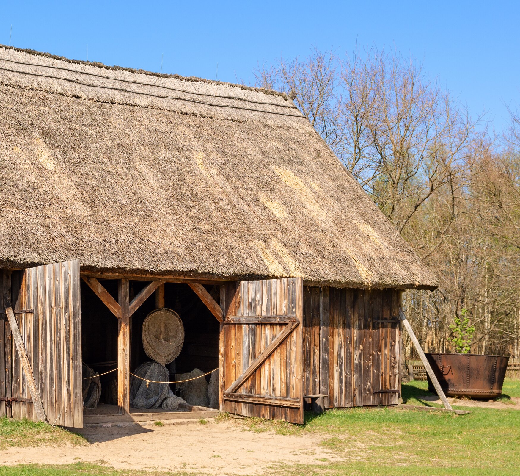 Kliknij, aby powiększyć. Zdjęcie przedstawia fragment drewnianej szopy. Jest jednopiętrowa i posiada dwuspadowy dach pokryty trzciną. Wokół zagrody znajduje się trawnik.