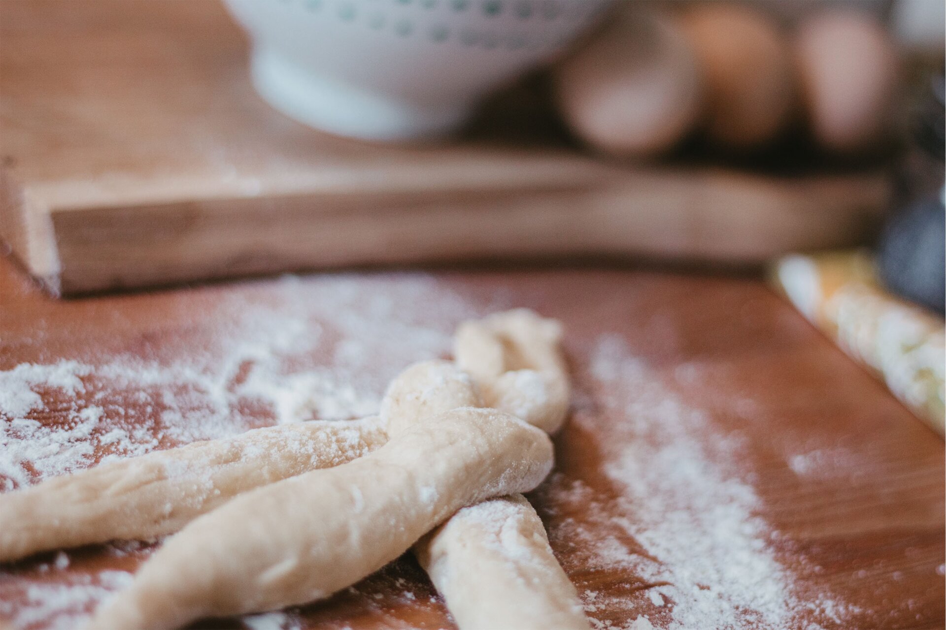 Zdjęcie przedstawia krok czwarty formowania chały tureckiej. Ujęcie wykonane jest od góry pod skosem. Widoczny jest ciemnobrązowy blat obsypany białą mąką. Na pierwszym planie w dolnej lewej części grafiki znajdują się trzy podłużne owalne wałki ciasta oprószone mąką. Końce wałków z jednej strony są ze sobą złączone, a w pozostałej części długości wałki wzajemnie się oplatają. Na drugim planie dostrzec można niewyraźny zarys jasnobrązowej drewnianej deski na której stoi biała miseczka, a obok po prawej stronie widoczne są trzy jajka. W prawej części grafiki niewyraźnie widoczny jest fragment białego obrusu w zielone i żółte motywy roślinne.