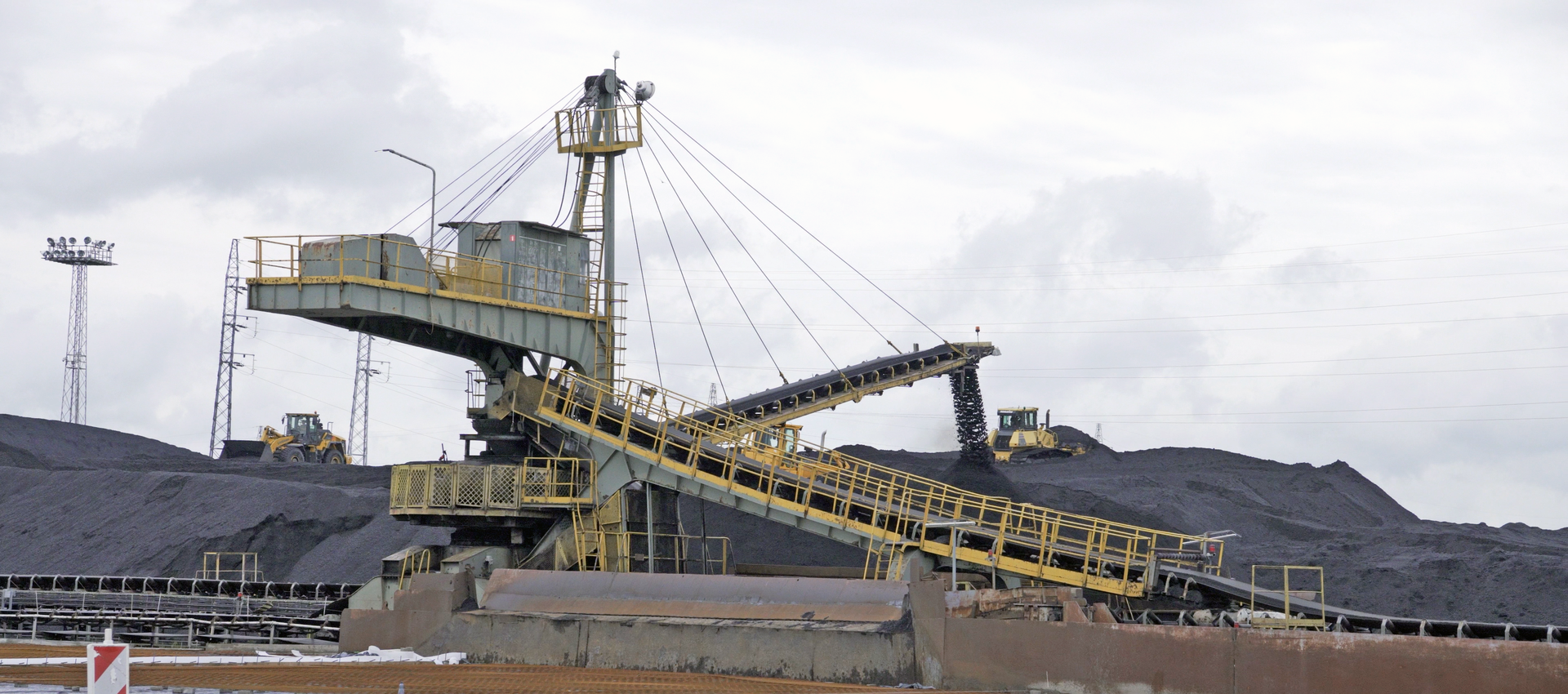 Na zdjęciu widoczna jest zwałowarka. Zwałowarka jest maszyną wielkogabarytową. Z prawej strony do zwałowarki dołączony jest przenośnik taśmowy. Z przodu zwałowarki znajduje się drugi przenośnik taśmowy. Przenośnik jest odchylony od poziomu. Przenośnik podwieszony jest na linach. Liny zamocowane są na głównym przęśle. Z przenośnika spada zrzucany na hałdę węgiel. W tle widać dużą hałdę węgla po której jeździ żółta ładowarka.