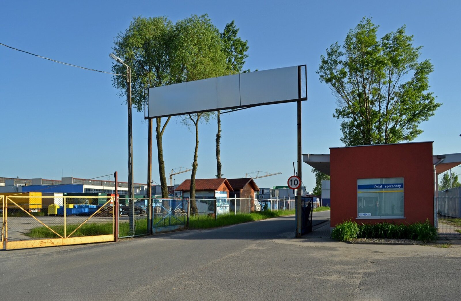 Wąska droga asfaltowa prowadząca do ogrodzonego terenu przemysłowego z halami. Drogę przedziela szlaban, znak ograniczenia prędkości do dziesięciu kilometrów na godzinę. Nad szlabanem poziomy baner z nazwą i logo strefy Interior‑Zachód oraz adresem ulica Jerzmanowska 8.