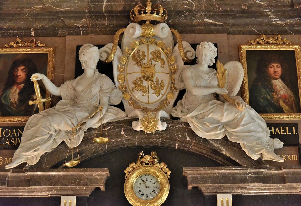 Fotografia przedstawiająca rzeźbę Alegorii Sprawiedliwości i Pokoju. Rzeźba przedstawia anioła i kobietę z mieczem i wagą, dookoła nich widoczne są złote wykończenia i marmur. Między kobietą a aniołem umieszczona jest owalna tarcza zwieńczona koroną. Na tarczy widnieją naprzemiennie sylwetki orła w koronie i Pogoni Litewskiej. Obok rzeźby wiszą na ścianach portrety mężczyzn, poniżej okrągły, złoty zegar.