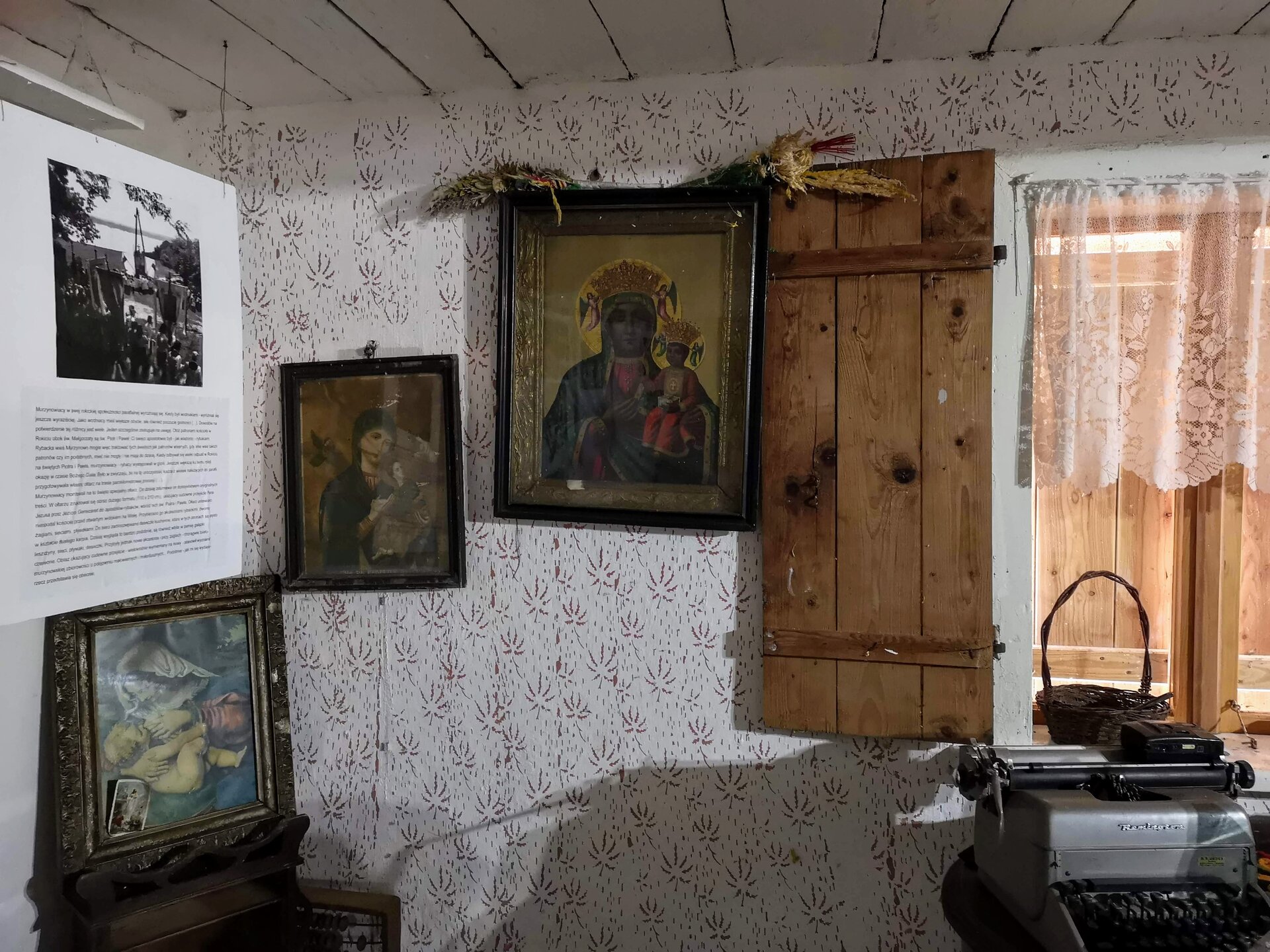 Fotografia przedstawia wnętrze starej chaty. Na niej po prawej stronie drewniana okiennica obok okna, po lewej stronie na wzorzastej ścianie trzy ułożone na skos obrazy świętej rodziny.